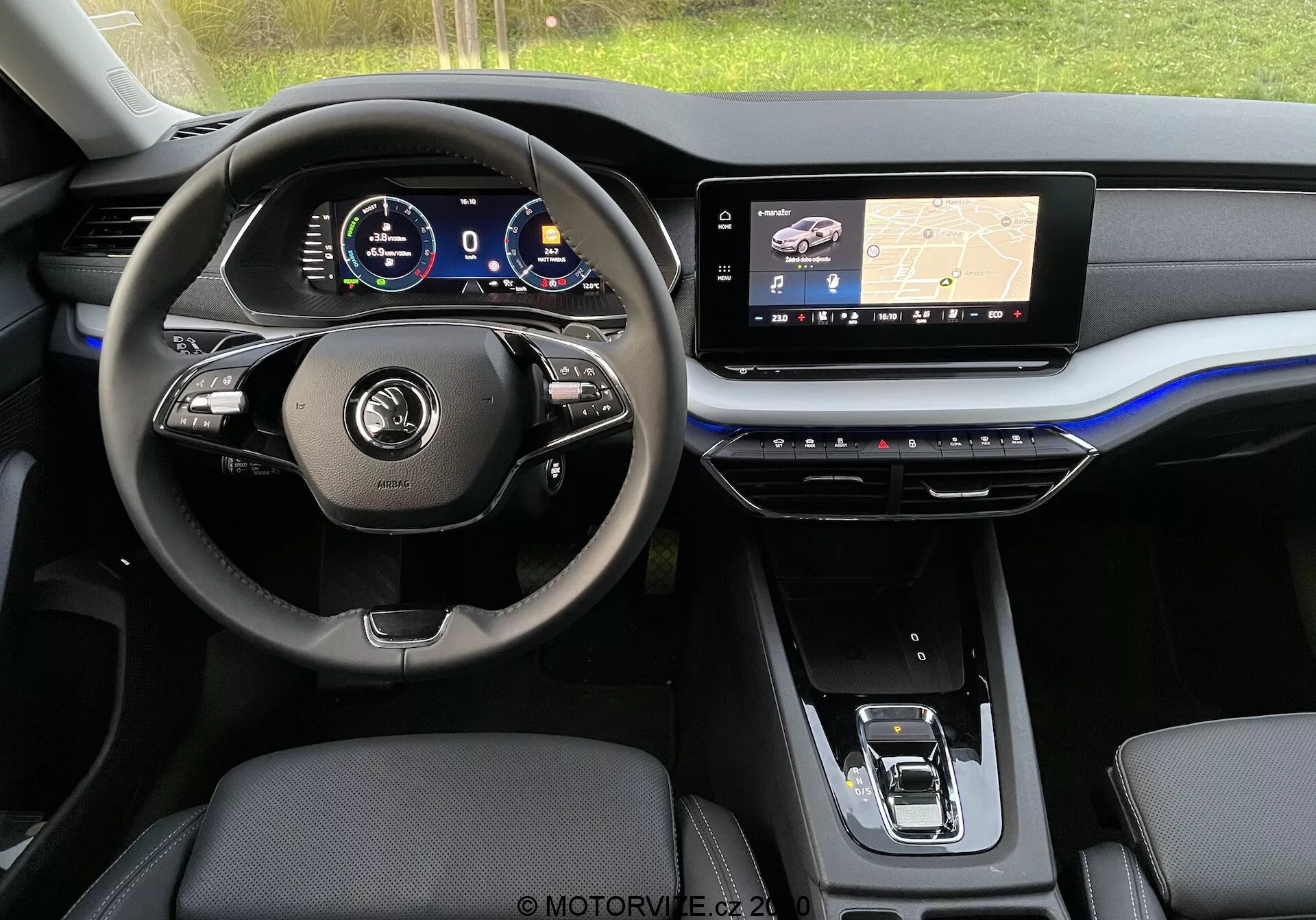 Pohľad na interiér moderného vozidla Škoda Octavia Limousine 2020 zo zadného sedadla, zobrazujúci prednú palubnú dosku so zameraním na oblasť vodičovho sedadla. Volant má multifunkčné tlačidlá a kožený poťah, za ním je digitálny prístrojový panel zobrazujúci rôzne ukazovatele vozidla. Stredná palubná doska obsahuje veľký infotainment dotykový displej zobrazujúci navigačnú mapu s GPS technológiou, zatiaľ čo stredová konzola nižšie obsahuje dotykové tlačidlá na ovládanie klímy. Viditeľné sú predné sedadlá s koženým poťahom a elektronický volič prevodových stupňov. Estetika je elegantná s tmavými tónmi, kovovými akcentmi a odvážnym modrým zvýraznením cez palubnú dosku.