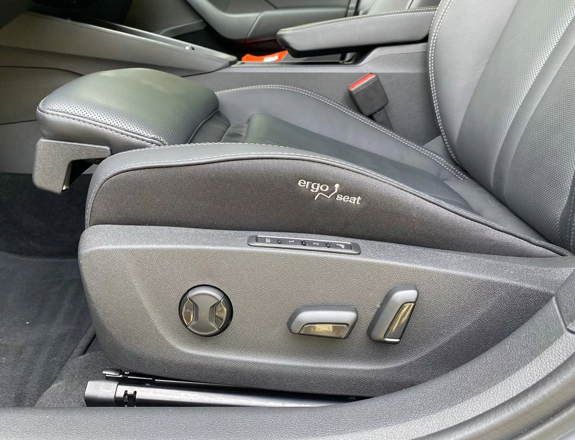 Detailný pohľad na bočné ovládanie sedadla vodiča vo vozidle Škoda Octavia 2020, zobrazujúci ergonomický dizajn s tmavým látkovým poťahom, viditeľným štítkom 'ergo seat' a elektronickými nastavovacími prvkami, vrátane kruhového naklápacieho ovládača a dvoch kovových tlačidiel na polohovanie, zdôrazňujúci prémiový a technologicky pokročilý interiér vozidla.