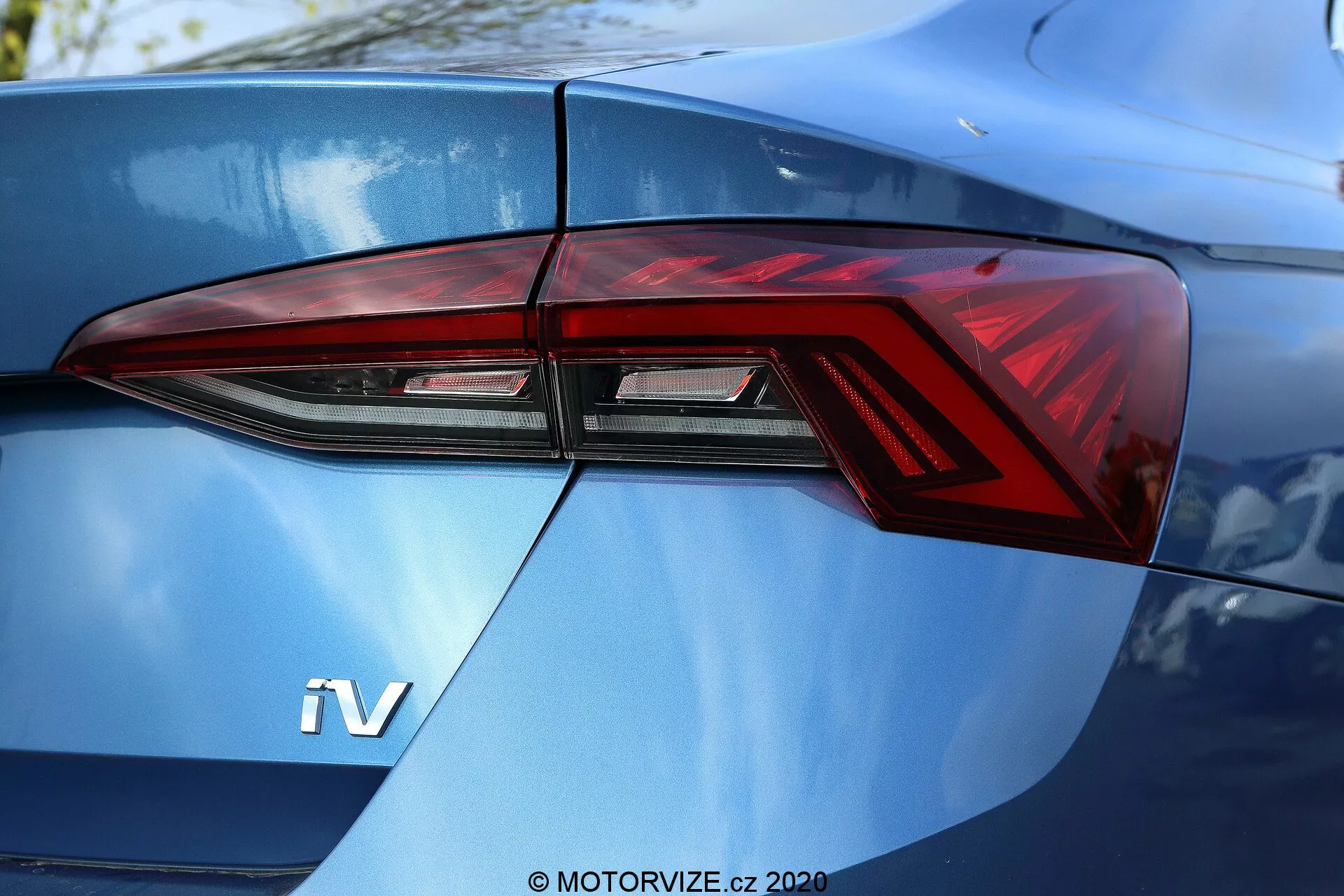 Detailný záber na zadnú časť vozidla Škoda Octavia Limousine 2020, zameraný predovšetkým na zadné svetlo na ľavej strane, viditeľné z uhla tri štvrtiny. Moderný dizajn zadného svetla so strmými uhlami a výraznými LED prvkami, plynulo integrovanými do línií karosérie vozidla. Viditeľný odznak 'iV' na lesklom modrom kufri naznačuje hybridné alebo elektrické vozidlo.