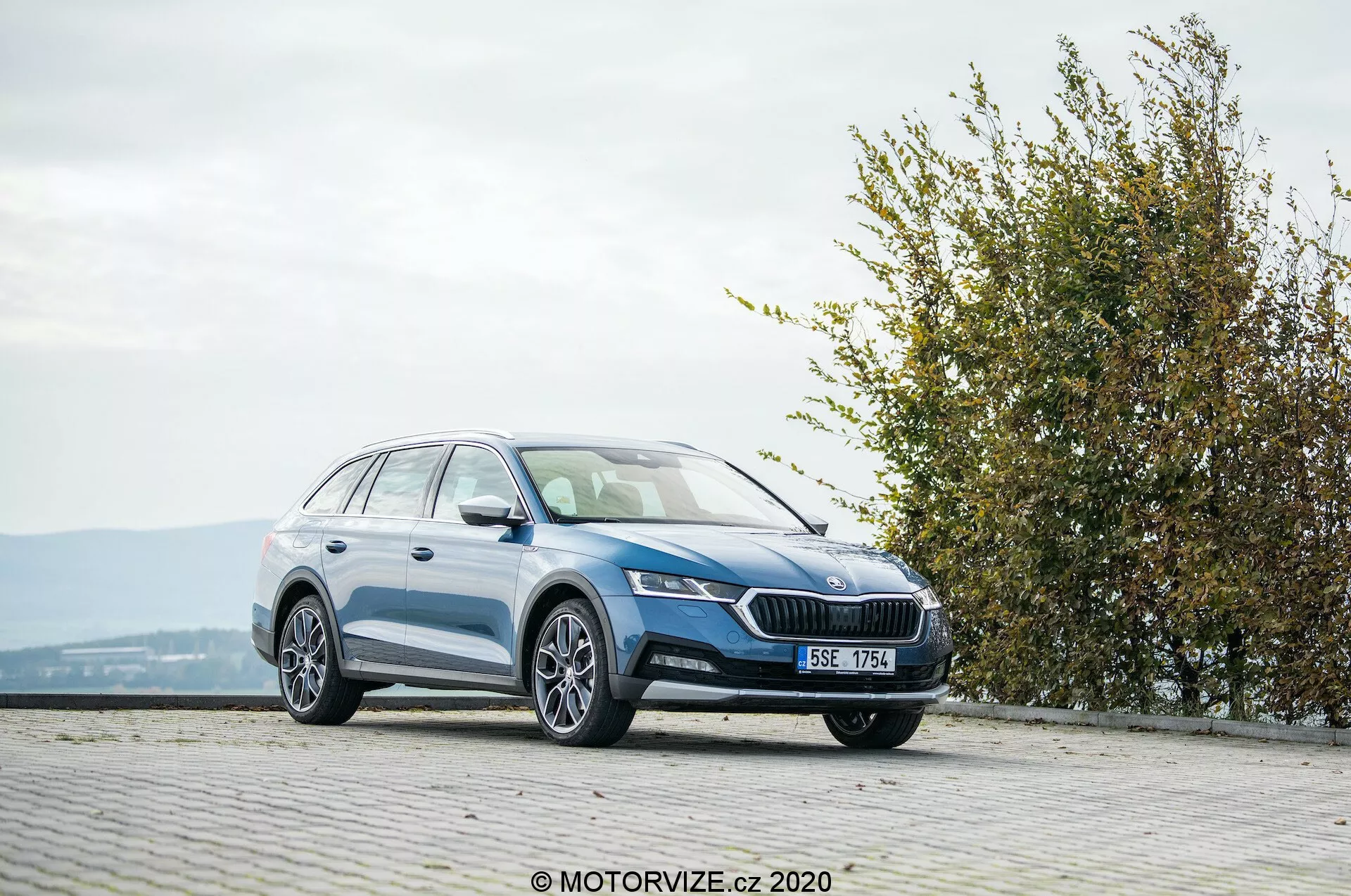 Veľký kombi Skoda Octavia Scout 2020, fotografovaný z trojštvrtinového predného pohľadu na dlhovanenej oblasti s kockami na vyvýšenom mieste. Obrázok zobrazuje prednú mriežku s výrazným emblémom, elegantné, uhlovité svetlá, integrované hmlovky, hladký bočný profil s hliníkovými kolesami, strešné nosiče a lesklý povrch laku. Okolie zahŕňa živý plot alebo krík vpravo, čo naznačuje blízkosť parku alebo zelenej plochy, v priaznivých poveternostných podmienkach.