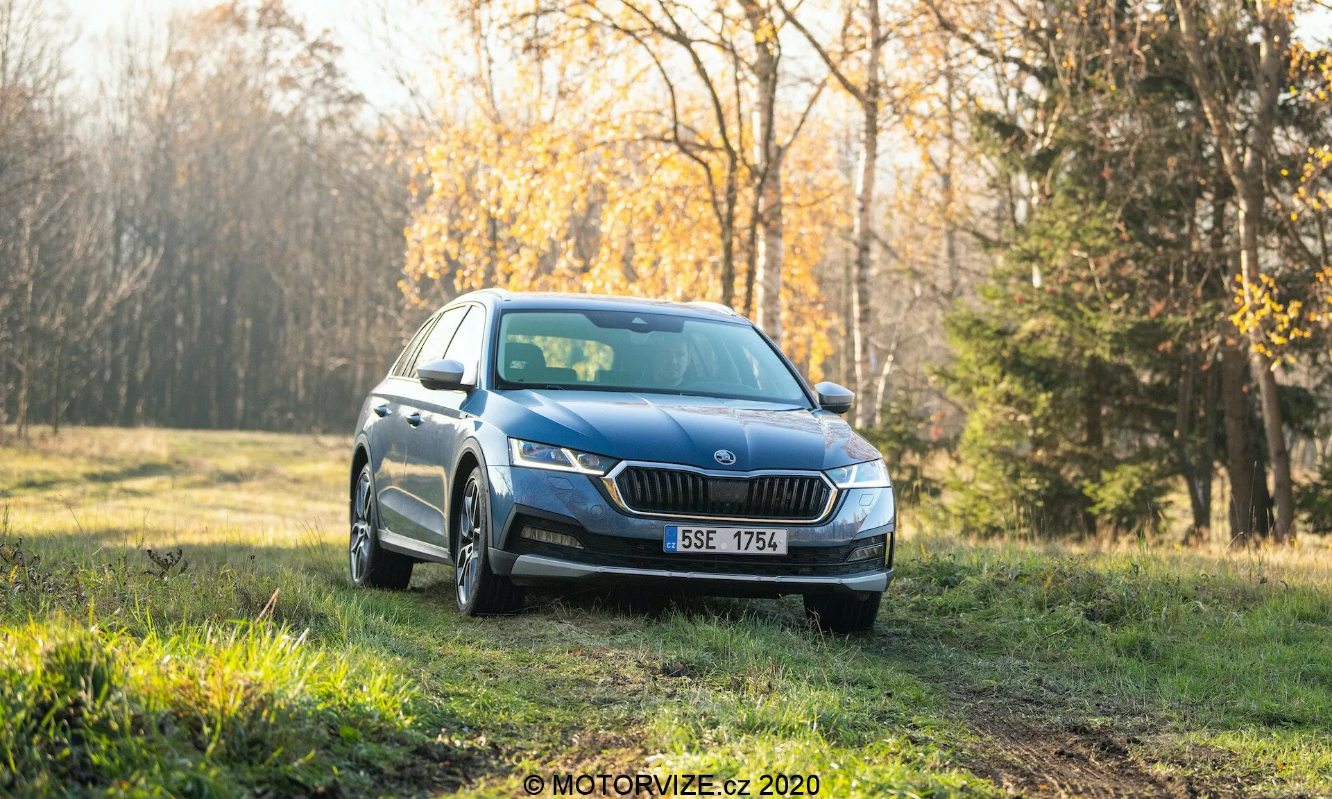 Modrý sedan Skoda Octavia Scout z roku 2020, fotografovaný z predného trojštvrťového uhla, zaparkovaný na trávnatej ploche s lesom v pozadí. Obrázok ukazuje moderné LED svetlomety, výraznú prednú masku, hmlové svetlá, viacramenné kolesá a integrované smerovky v bočných zrkadlách. Nastavenie vyzerá ako skoro ráno alebo neskoro popoludní s jemným slnečným svetlom a teplým tónom čistého auta určeného pre reklamu alebo recenziu. Rozmazané pozadie zvýrazňuje dizajnové prvky a pôsobivé vonkajšie prostredie vozidla.
