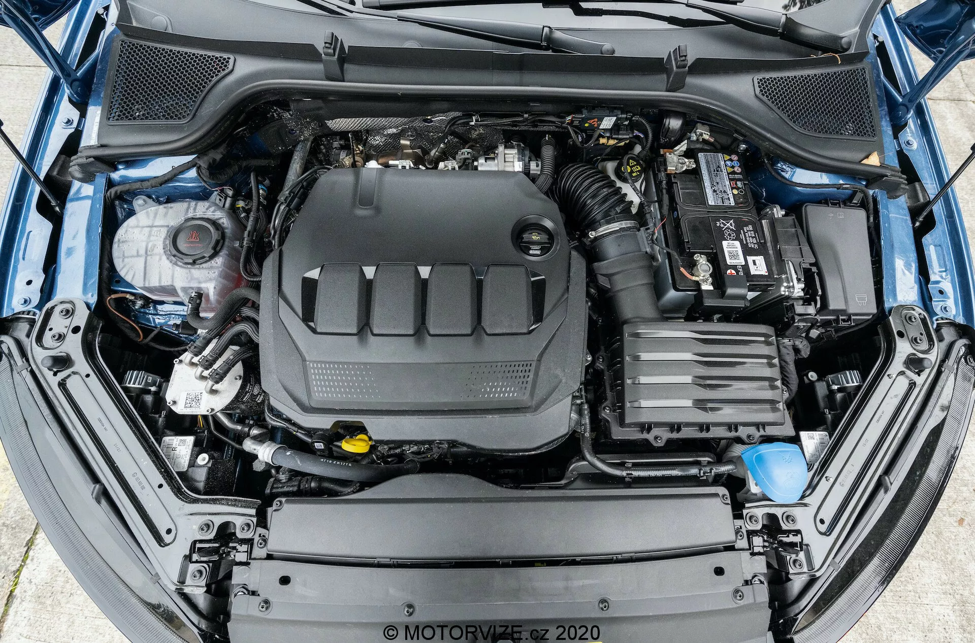 Pohľad z vtáčej perspektívy na otvorenú motorovú časť automobilu, ktorý ukazuje komplexné usporiadanie motora a jeho okolitých komponentov. Motorový blok je centrálne umiestnený a zakrytý plastovým krytom motora. Napravo je viditeľná batéria automobilu s viditeľnými pripojeniami na hornom konci. Predná časť zobrazuje komponenty systému prívodu vzduchu. Vľavo sú rôzne nádrže na kvapaliny, vrátane nádržky na brzdovú kvapalinu so žltým viečkom. Viditeľné sú tiež rôzne hadice a kabeláž. Štruktúra predného nárazníka a mriežky je čiastočne viditeľná na dolnom okraji obrázka.