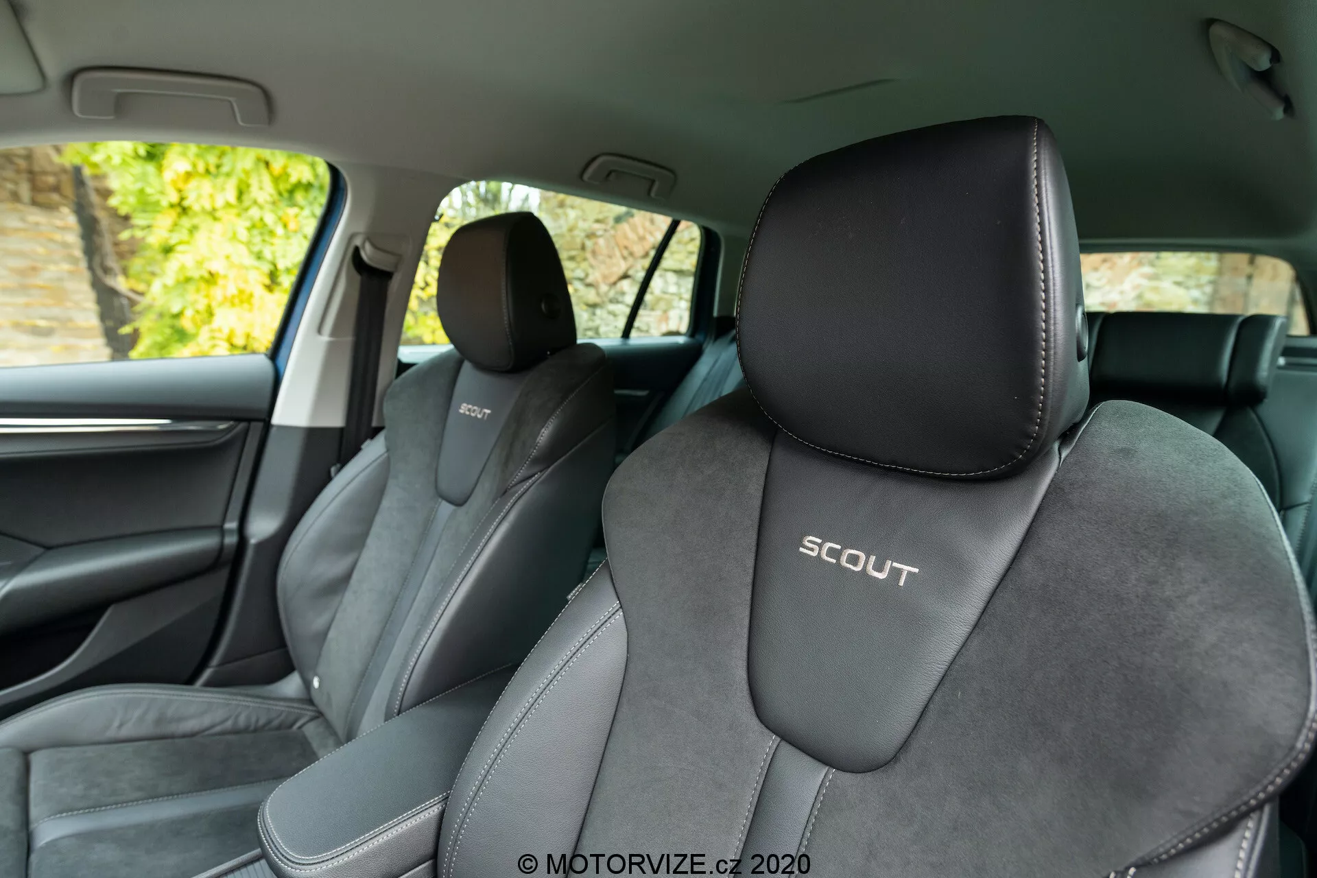 Pohľad na interiér Škoda Octavia Scout 2020 z zadných sedadiel, ktorý ukazuje predné sedadlá. Uhol favorizuje ľavú stranu, čo poskytuje jasný pohľad na vodičovo sedadlo s embosovaným logom 'SCOUT'. Sedadlá sú čalúnené dvojfarebne, prevažne čierne s svetlejšími šedými akcentmi a centrálnym materiálom zo semiše, čo zdôrazňuje športový dizajn. Obrázok zachytáva aj B-stĺpik, tmavé interiérové lišty, časť predných dverí, mriežku reproduktora a chrómovú kľučku dverí. Pozadie za oknami naznačuje lístie, čo ukazuje na zaparkované auto vonku v prirodzenom svetle.