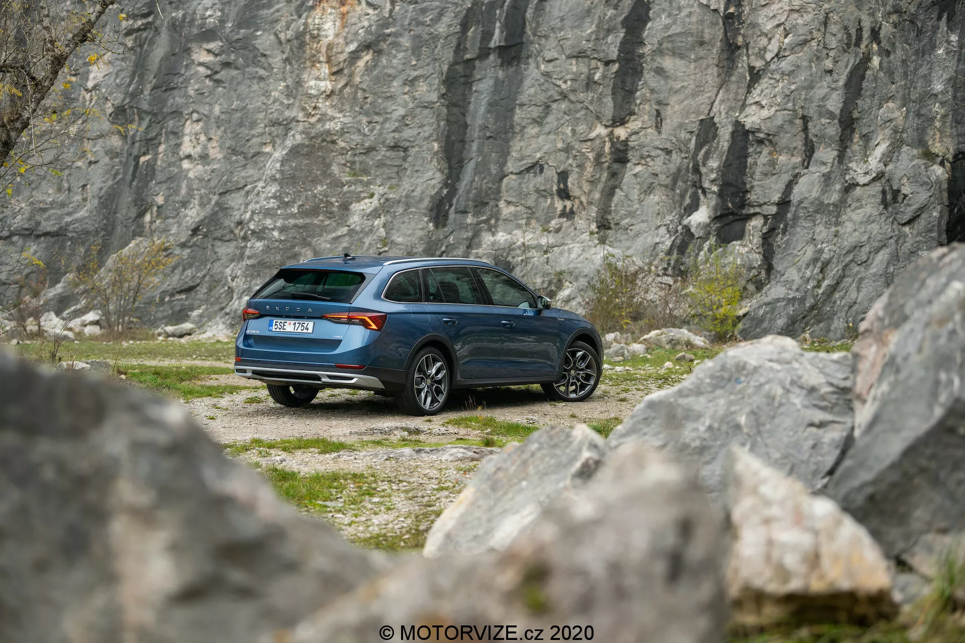 Obrázok zobrazuje auto Skoda Octavia Scout 2020 zaparkované uprostred skalnatého terénu s kameňolomom alebo útesom v pozadí, zachytené z mierneho teleobjektívového pohľadu. Trojštvrťový zadný uhol poskytuje podrobný pohľad na bočný profil a zadnú časť vozidla, ktorý ukazuje moderný dizajn kombi, šikmú zadnú strechu a tmavomodrú farbu. Viditeľné prvky zahŕňajú zadné svetlá, tvar okien, zadný stierač, dizajn kolies a bočné zrkadlá. Prostredie evokuje tému dobrodružstva a prieskumu, čo naznačuje vhodnosť vozidla pre jazdu v ťažko dostupných miestach.