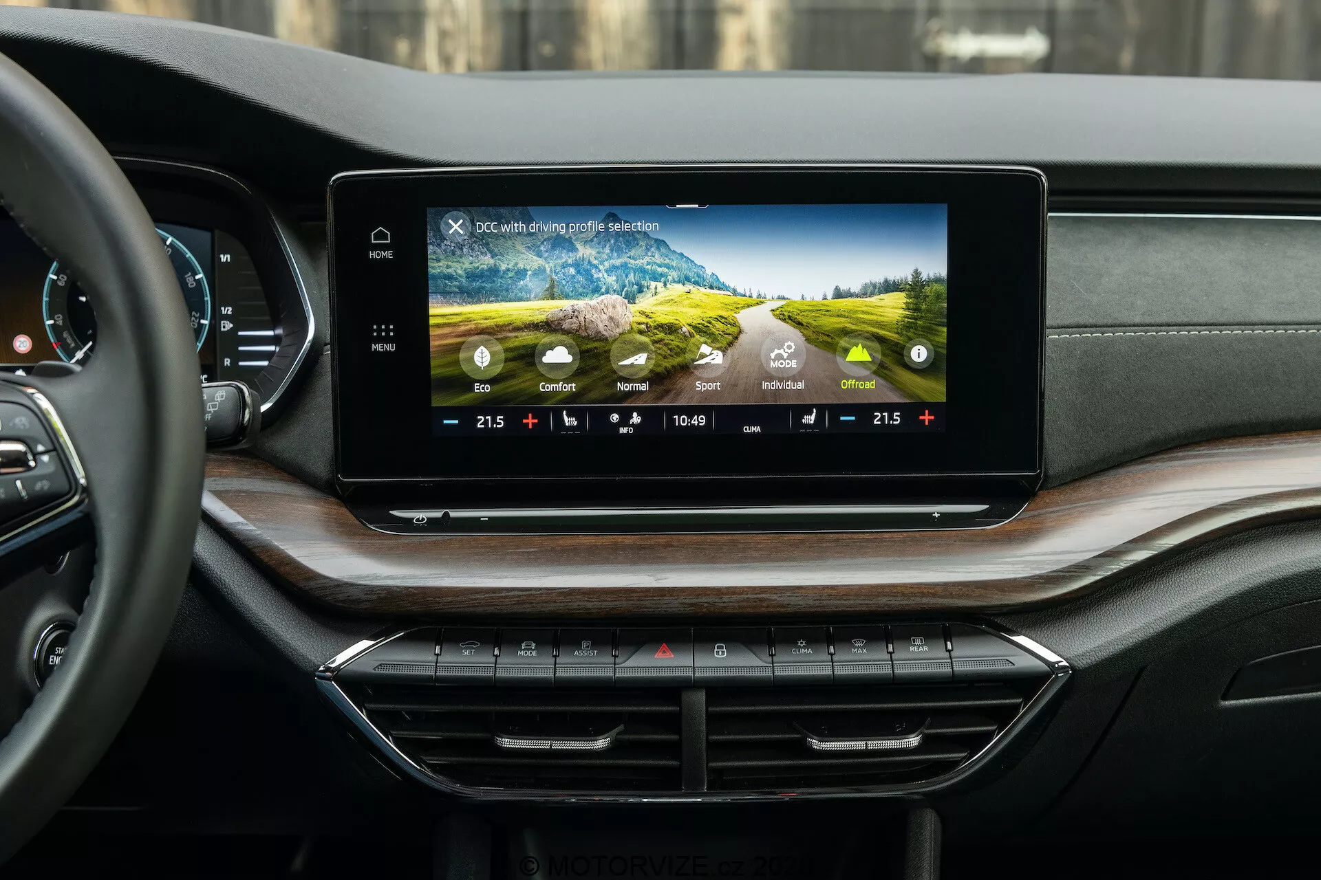 Pohľad na interiér vozidla Škoda Octavia 2020 (Octavia Scout) s moderným infotainment systémom a prístrojovou doskou, fotografovaný z perspektívy zadného sedadla. Na palubnej doske je široká horizontálne orientovaná dotyková obrazovka zobrazujúca farebné grafické rozhranie, nastavenia klimatizácie, čas a vonkajšiu teplotu. Pod nimi sú úhľadne usporiadané tlačidlá a ovládače klimatizácie. Luxusné prístrojové doske dominuje mäkký materiál a drevená dyha, s diskrétne integrovanými vzduchovými otvormi.