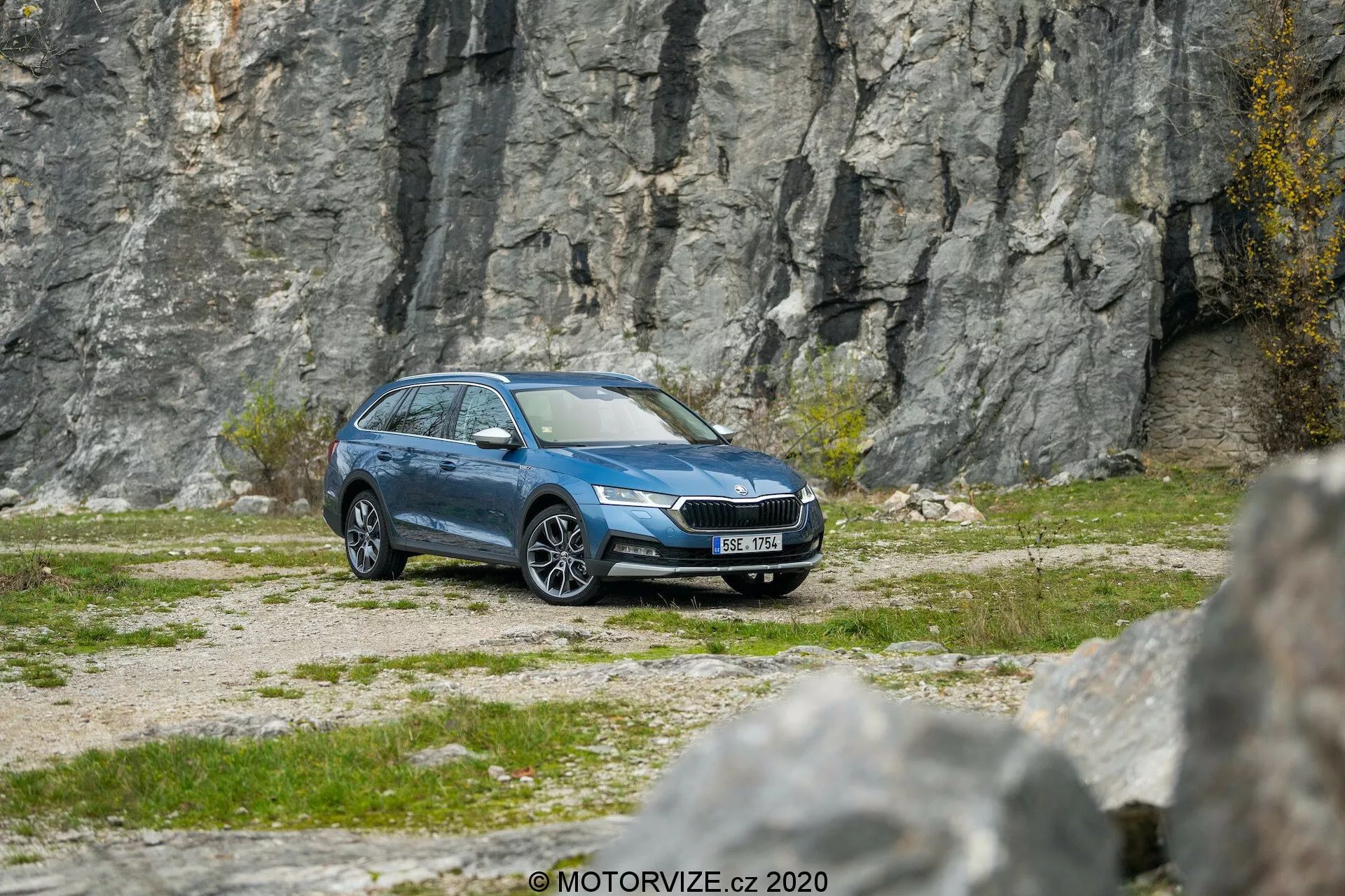 Bočný predný pohľad na modrú Škoda Octavia Scout 2020 kombi na štrkovom teréne so skalnatým pozadím, zobrazujúci moderné LED svetlomety, výraznú prednú mriežku, hliníkové kolesá, bočné zrkadlá, okenné lišty a strešné nosiče a čistý metalický povrch, naznačujúci pokročilé funkcie ako technológia osvetlenia, monitorovanie tlaku v pneumatikách a možné bezpečnostné systémy.