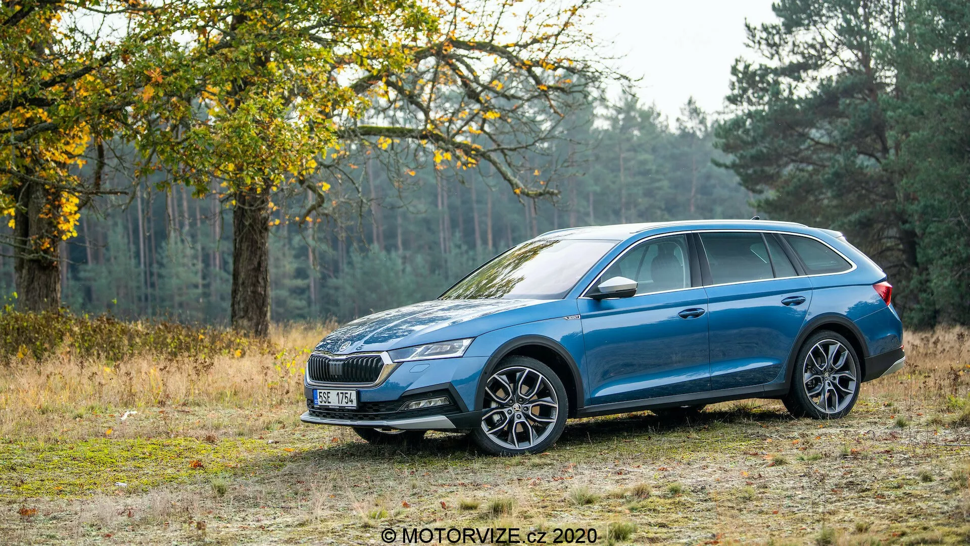 Bočný profil modrého kombi Skoda Octavia Scout 2020 na prašnej ceste s pozadím stromov a trávy, ukazujúci jeho schopnosť zvládať terénne podmienky. Auto má metalický lak, viac-lúčový dizajn hliníkových kolies, LED svetlomety, výraznú prednú mriežku, strešné nosiče, zrkadlá a kľučky dverí v farbe karosérie a strešné okno. Dizajn zahŕňa charakterovú líniu, ktorá sa tiahne od predného oblúku kolesa po zadné svetlá a obklopujúce zadné svetlá.