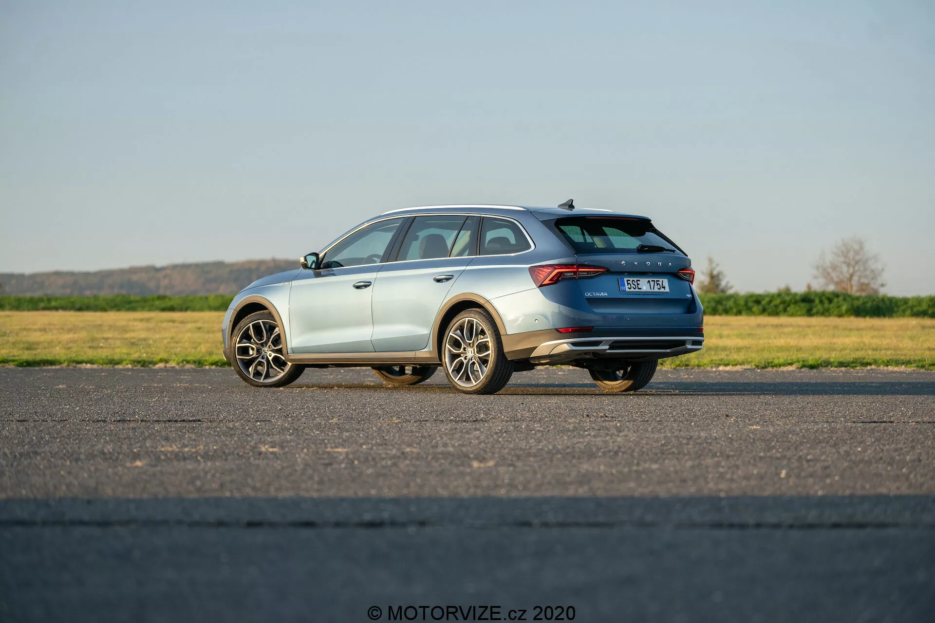 Obrázok ukazuje Škodu Octavia Scout 2020, moderné kombi, zachytené z trojštvrťového zadného pohľadu. Zvýrazňuje prvky dizajnu vozidla vrátane horizontálnych zadných svetiel, ktoré sa tiahnu k stredu zadných dverí, strešného spojlera, strešných nosičov a viacramenných zliatinových kolies. Vozidlo je umiestnené na rovnom dláždenom povrchu, pričom bočná strana vodiča je bližšie k fotoaparátu. Vozidlo má metalický lak a časť výfukového systému. EČV je mierne rozmazaný a viditeľné je logo Škoda. Fotografia bola zhotovená za denného svetla pod jasnou oblohou a zobrazuje stojace vozidlo s možnými skrytými technologickými prvkami ako je parkovací asistent, zadné kamery alebo systémy prevencie kolízií.