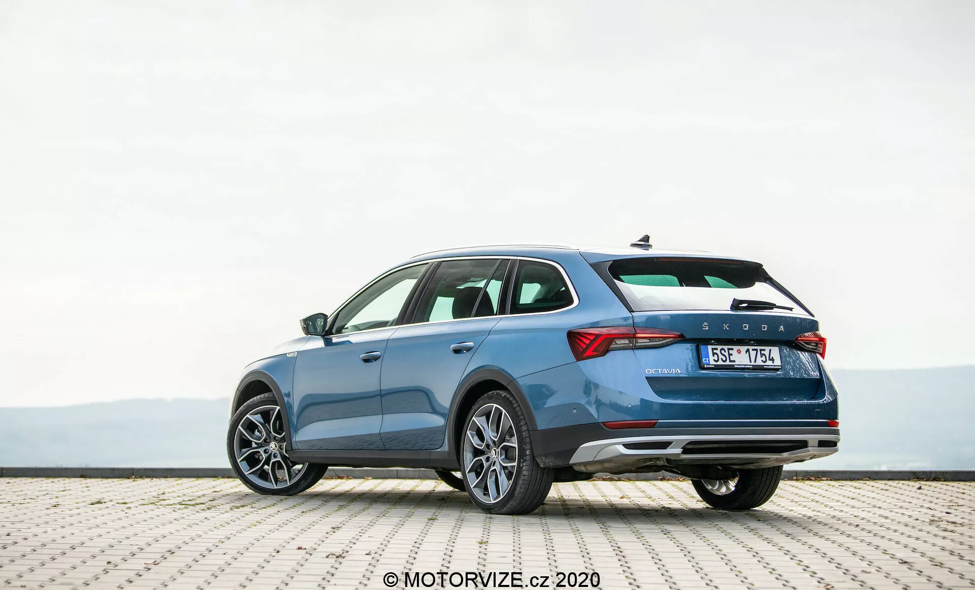 Podrobný záber na modré kombi Škoda Octavia Scout z roku 2020, zaparkované na dláždenom povrchu, zachytené z trojštvrťového zadného pohľadu. Fotografia zdôrazňuje elegantný profil vozidla, charakteristický dizajn zadných dverí a pravú stranu. Viditeľné prvky zahŕňajú horizontálne zadné svetlá, líniu karosérie naznačujúcu pohyb, zadný nárazník s odrazkami, ktoré môžu obsahovať parkovacie senzory, dvojitý výfukový systém a viaclúčové hliníkové kolesá. Obrázok naznačuje priestranný interiér, pokročilé bezpečnostné prvky a moderné dizajnové prvky vhodné pre nadšencov automobilovej technológie.