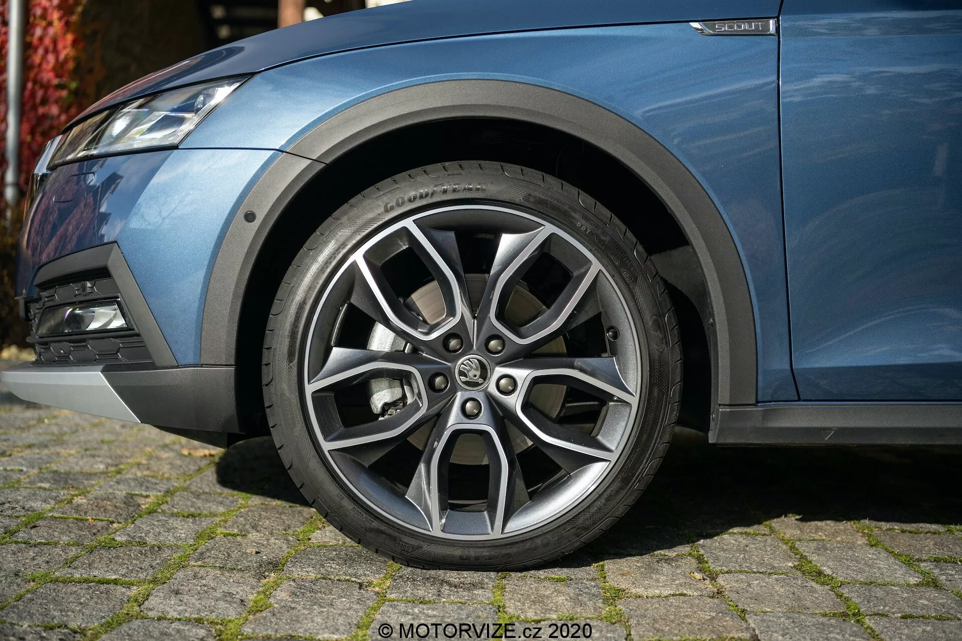 Detailný pohľad na prednú ľavú stranu vozidla Škoda Octavia 2020 (Octavia-Scout) so zameraním na starostlivo navrhnuté hliníkové koleso s viaclúčovým dizajnom, kontrastným strieborným povrchom a viditeľnými vzormi behúňa. Fotografia, urobená z nízkeho uhla, zachytáva časť kovového modrého tela vozidla, predný nárazník s hmlovkou a komponenty odpruženia, čo ukazuje moderný, športový vzhľad.