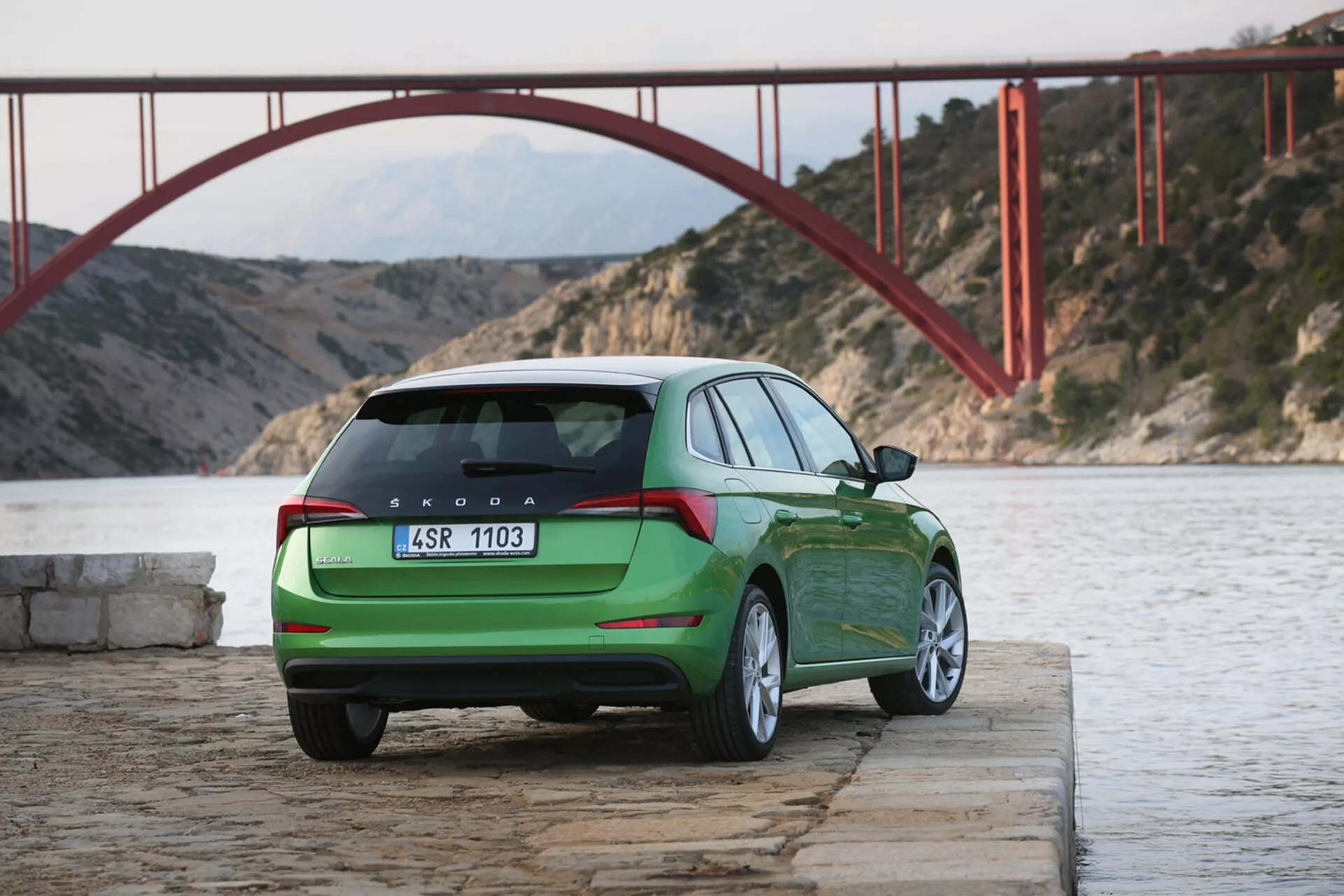 Zelená Škoda Scala z roku 2019 zaparkovaná pri vode s pohľadom na zadnú a bočnú časť, zobrazujúca zadné svetlá, zadné veko, logo a označenie modelu na pozadí s červeným mostom a horami