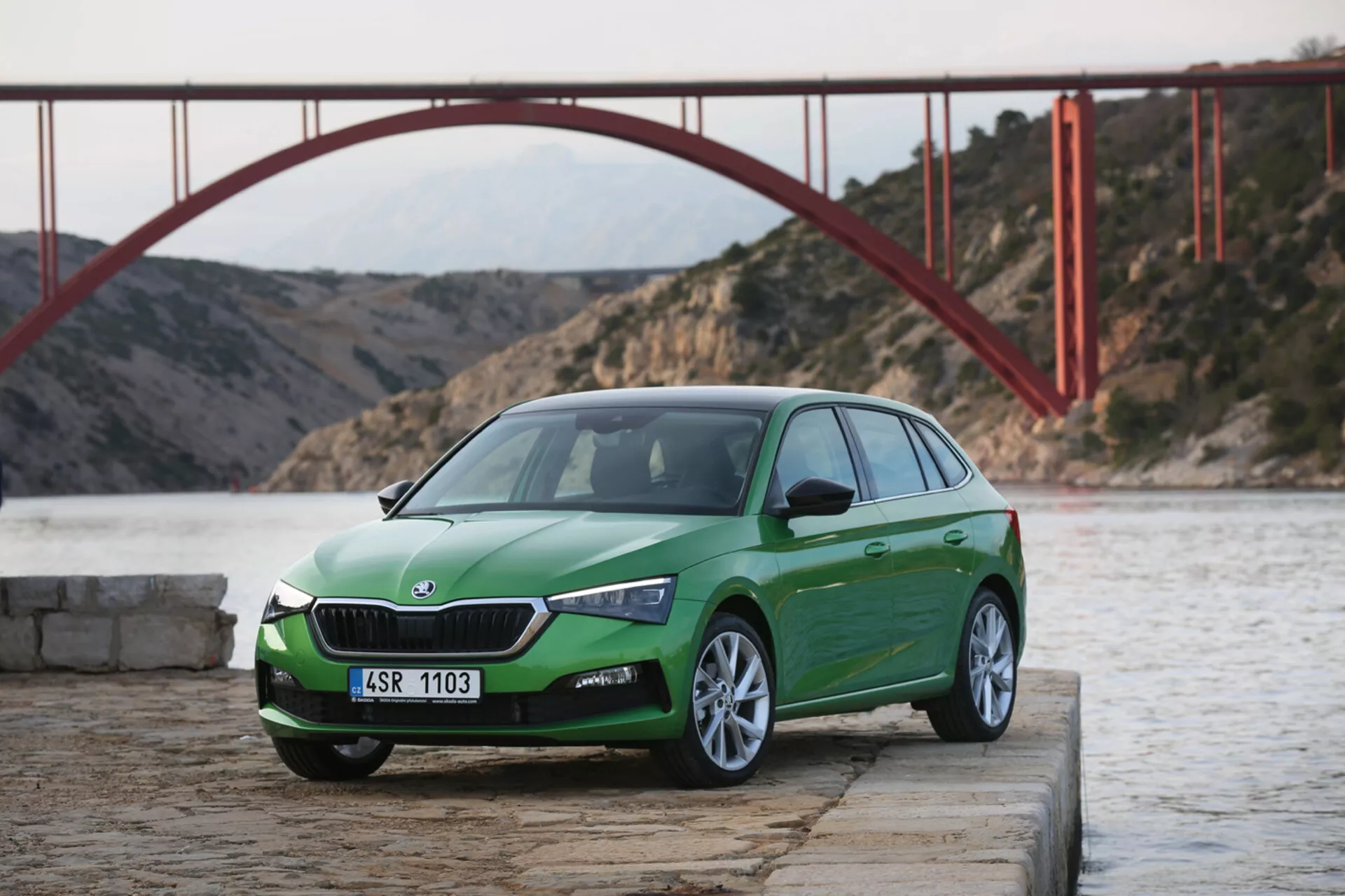 Uhlový predný trojštvrťový pohľad na zelený hatchback Skoda Scala 2019 na betóne pri vode s červeným mostom na pozadí počas dňa, predstavujúci dizajn a funkcie