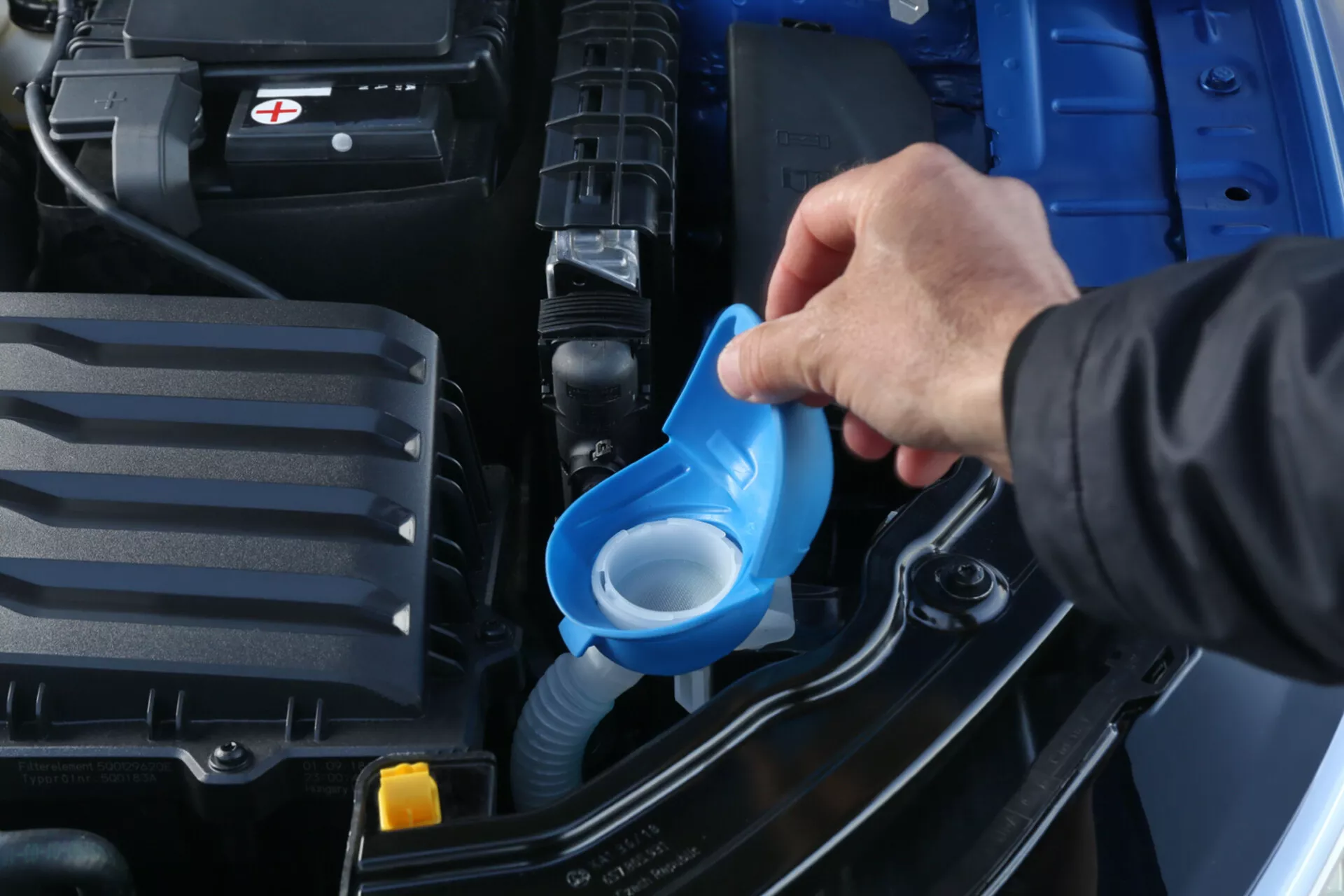 Detailný pohľad na motorový priestor Škoda Scala 2019 s dôrazom na dopĺňanie kvapaliny ostrekovača čelného skla pomocou modrého lievika, zobrazujúci batériu, kryt motora a konštrukciu vozidla