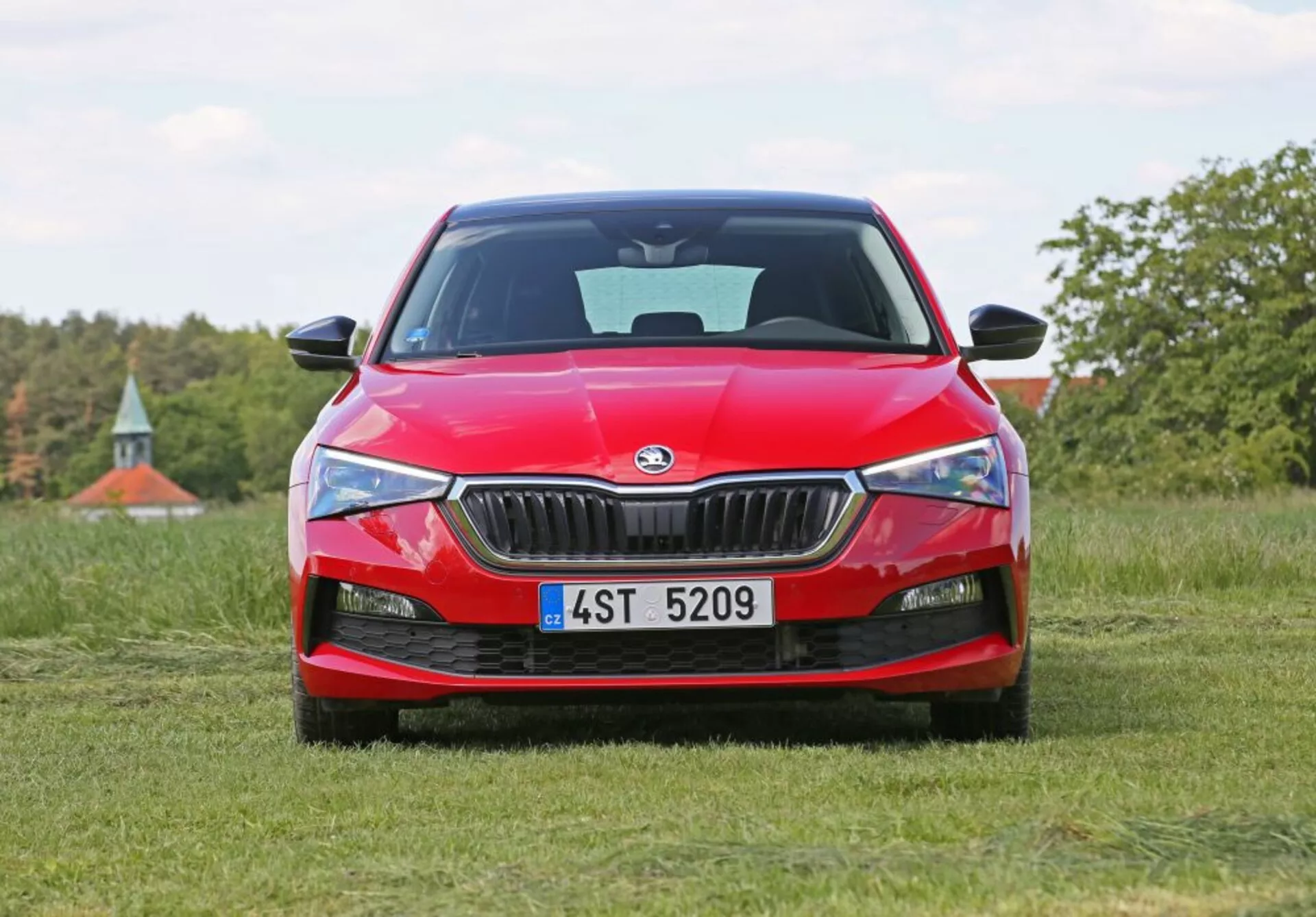 Škoda Scala 2019 v jasne červenej farbe, predstavujúca modernú technológiu LED svetlometov, uhlovú masku s chrómovým lemom a športový dizajn prednej časti, fotografovaná z nízkeho uhla na trávnom poli s prírodnou krajinou na pozadí