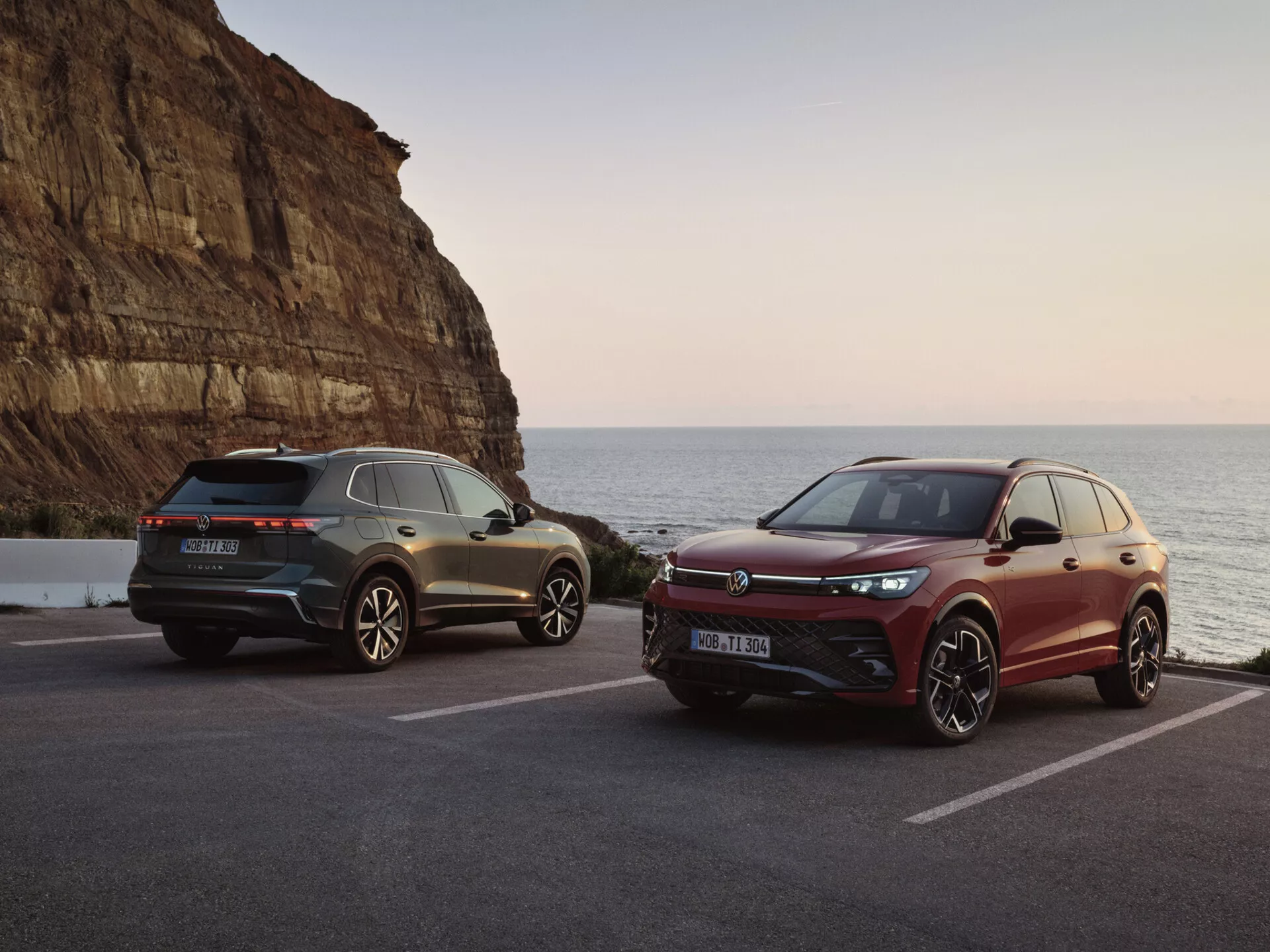 Luxusné SUV v malebnom pobrežnom prostredí počas západu slnka, so sivým modelom s LED zadnými svetlami a športovými dizajnovými prvkami a bordovým modelom s prednou mriežkou orientovanou na výkon a pokročilými systémami asistencie vodiča