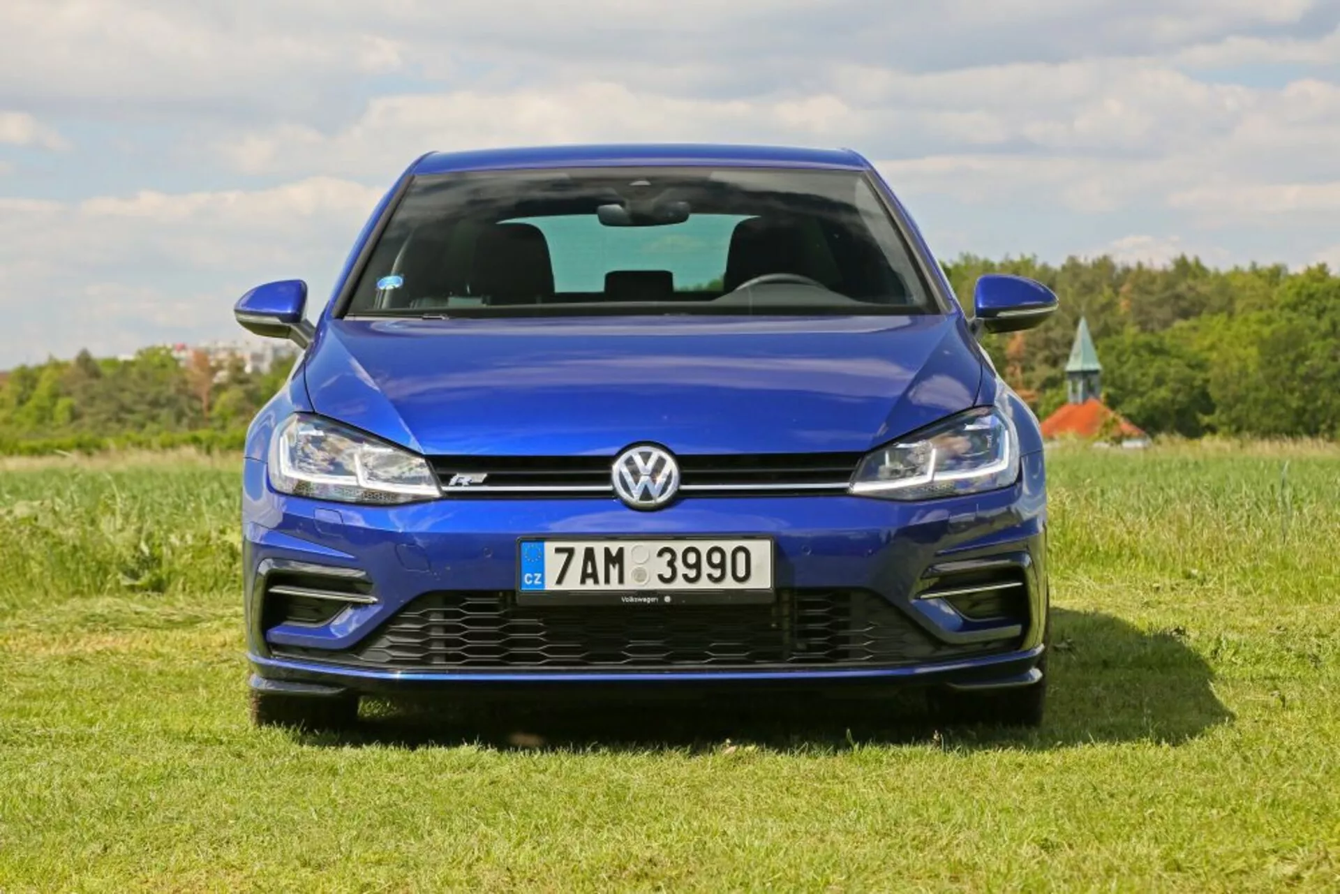Predný pohľad na modrý Volkswagen Golf zaparkovaný na trávnatom poli pod jasnou oblohou, zdôrazňujúci jeho kovový povrch, dizajn mriežky, technológiu LED svetlometov a športový predný nárazník vrátane nasávacích otvorov a emblému.