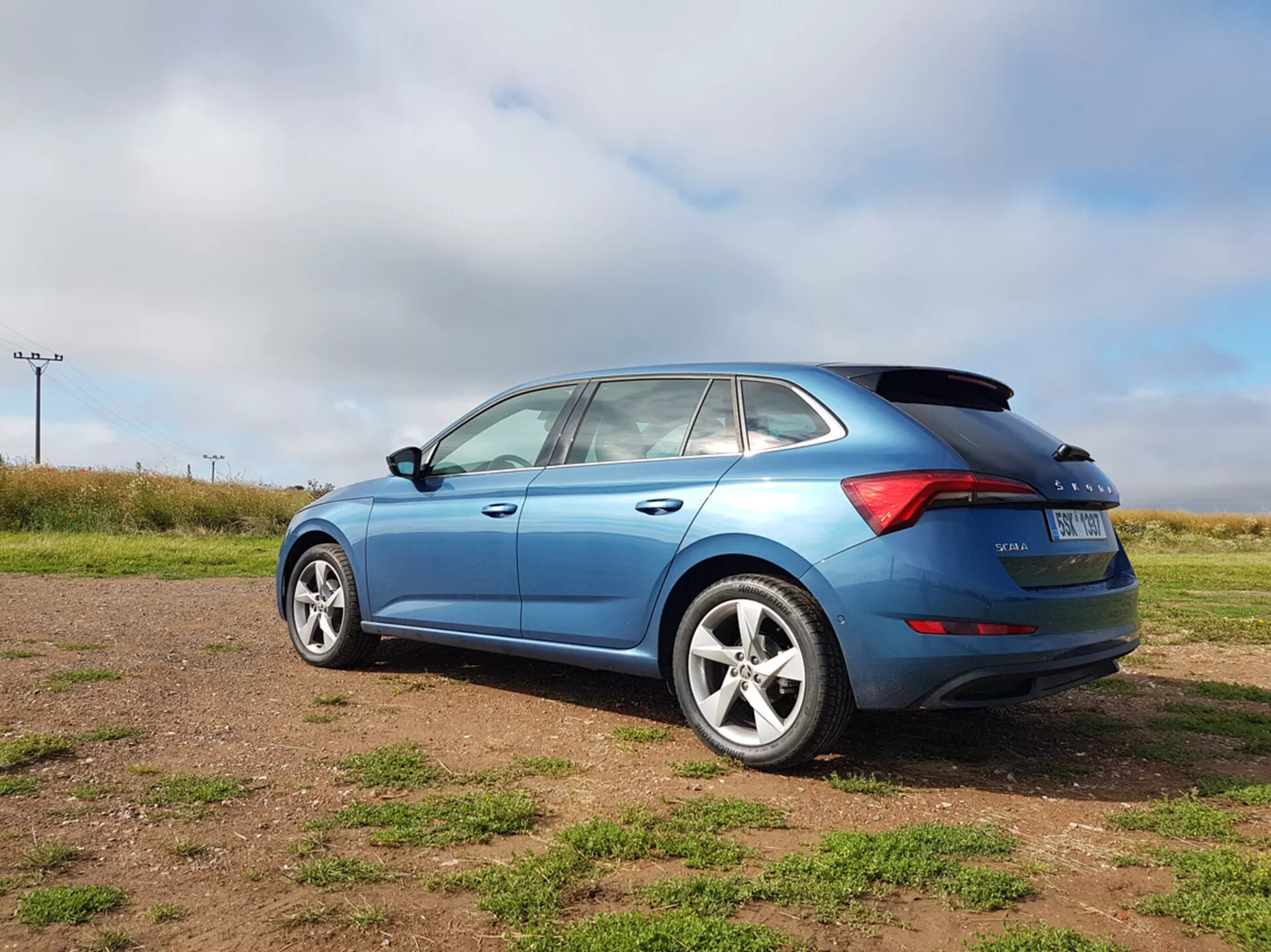 Modrý hatchback Skoda Scala z roku 2019 zobrazujúci bočné a zadné dizajnové prvky, vrátane tvaru okien, LED zadných svetiel a zliatinových kolies, fotografovaný z trojštvrťového zadného uhlu na trávnatom teréne