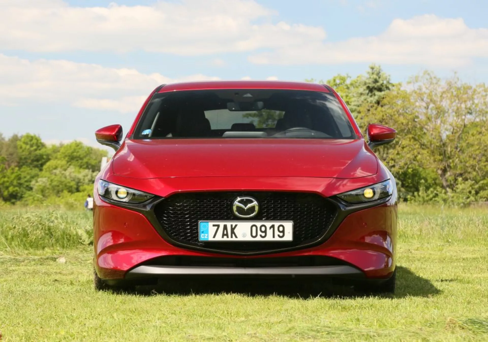 Vozidlo Mazda 2019 v červenej farbe, zobrazujúce čelný pohľad s výraznou mriežkou s emblémom Mazda, LED svetlometmi a profilovaným dizajnom kapoty. Umiestnené vonku na trávniku s pozadím stromov a modrej oblohy.
