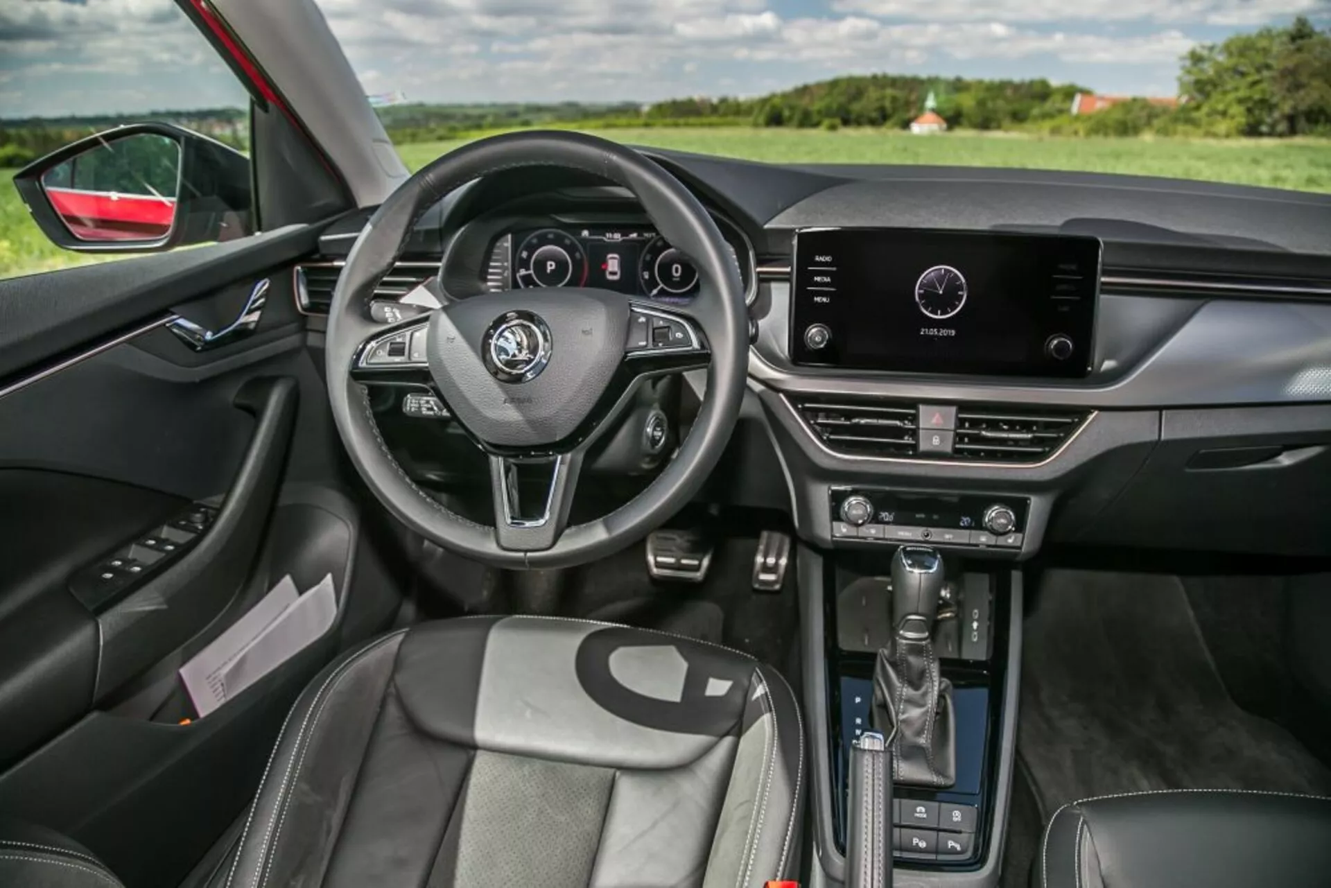 Interiér Škoda Scala 2019 z pohľadu spolujazdca s trojramenným volantom s ovládacími prvkami a digitálnym prístrojovým panelom, dotykovým infotainment systémom na hornej časti stredovej konzoly s manuálnymi ovládačmi klimatizácie nižšie, automatickou pákou prevodovky, profilovanými prednými sedadlami s koženkovým poťahom, dverovým panelom vodiča s elektrickými ovládaniami okien a zrkadiel, vnútorným ambientným osvetlením, zrkadlom s automatickým stmievaním a výhľadom na krajinu cez čelné sklo a okno vodiča, zdôrazňujúcim ergonomický dizajn a integrovanú technológiu.