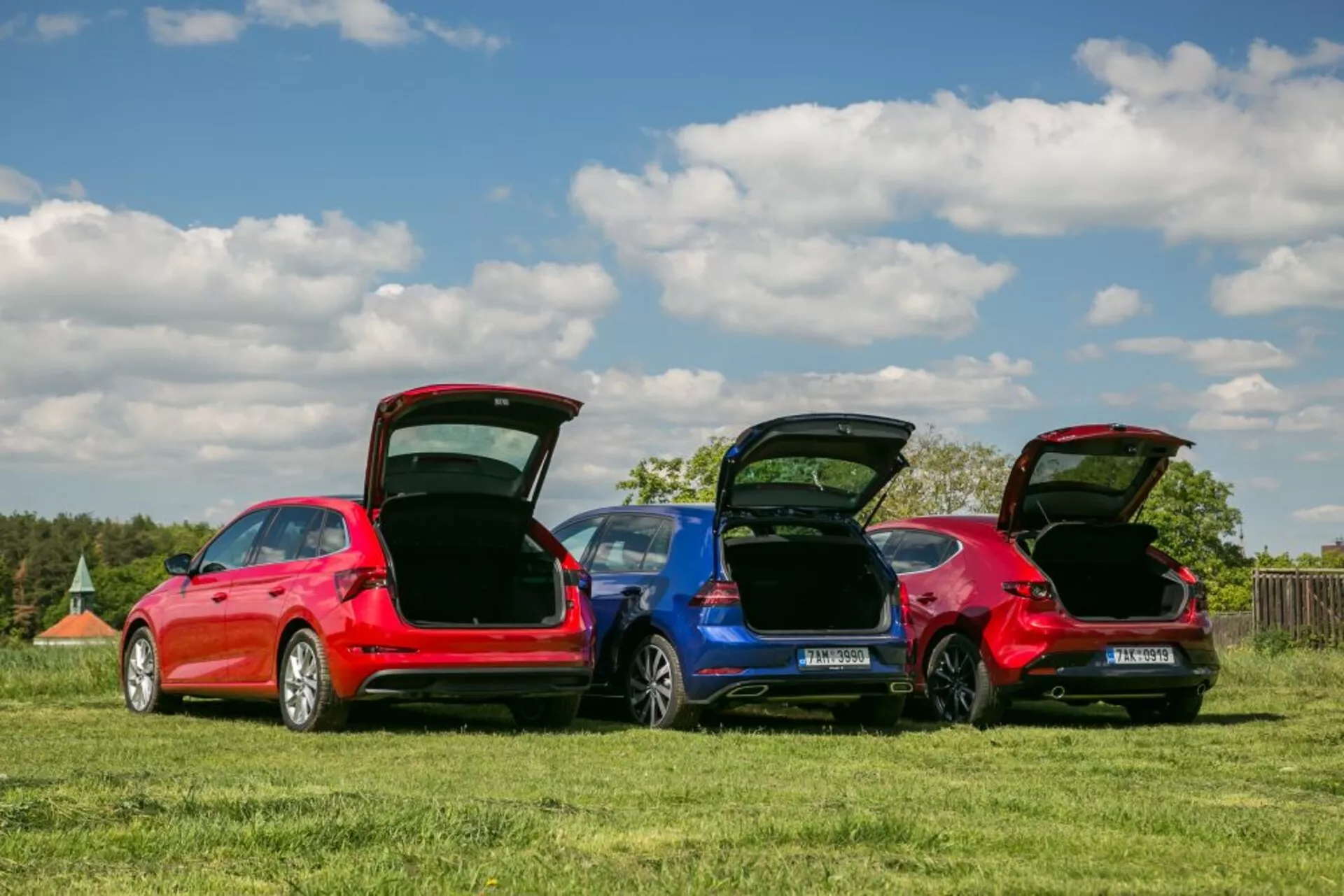 Modely Škoda Scala 2019 v červenej a modrej farbe zaparkované na trávnatom poli, ukazujúce zadné profily, otvorené batožinové priestory pre funkčnosť nákladového priestoru a detaily dizajnu pod jasnou oblohou.