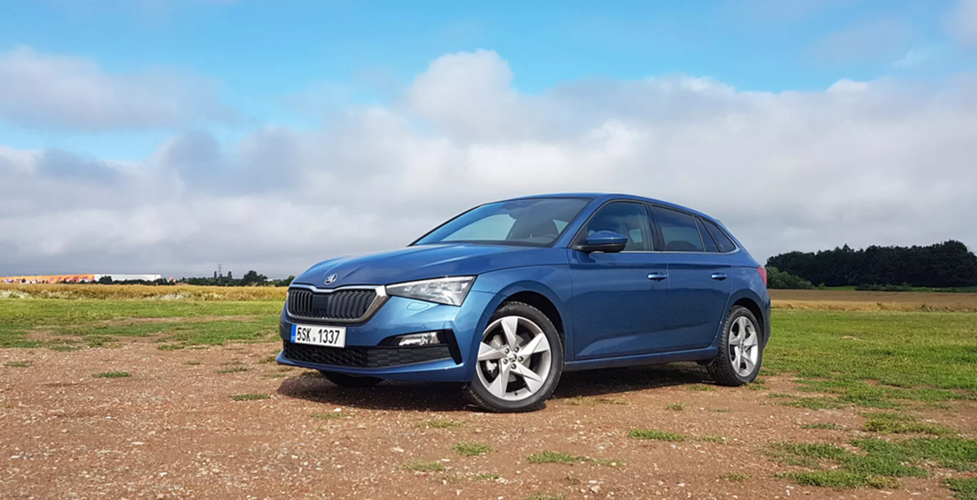 Modrý hatchback Škoda Scala 2019 na nezpevnenom povrchu s charakteristickou prednou mriežkou, modernými LED svetlometmi, zliatinovými diskami a elegantným profilom ľavej strany na pozadí čiastočne zamračenej oblohy