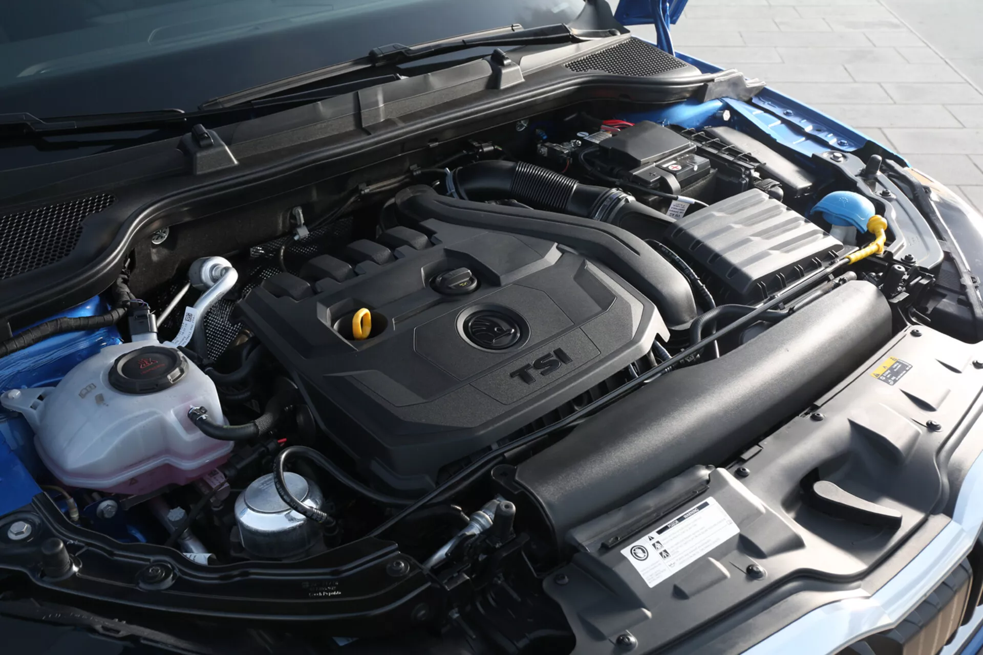 Detailný pohľad na motorový priestor Škoda Scala 2019 s dôrazom na kryt motora TSI s turbodúchadlom a komponenty vrátane nádrže na chladiacu kvapalinu, nádoby na ostrekovač, autobatériu a sústavu nasávania z mierne pravého predného uhla