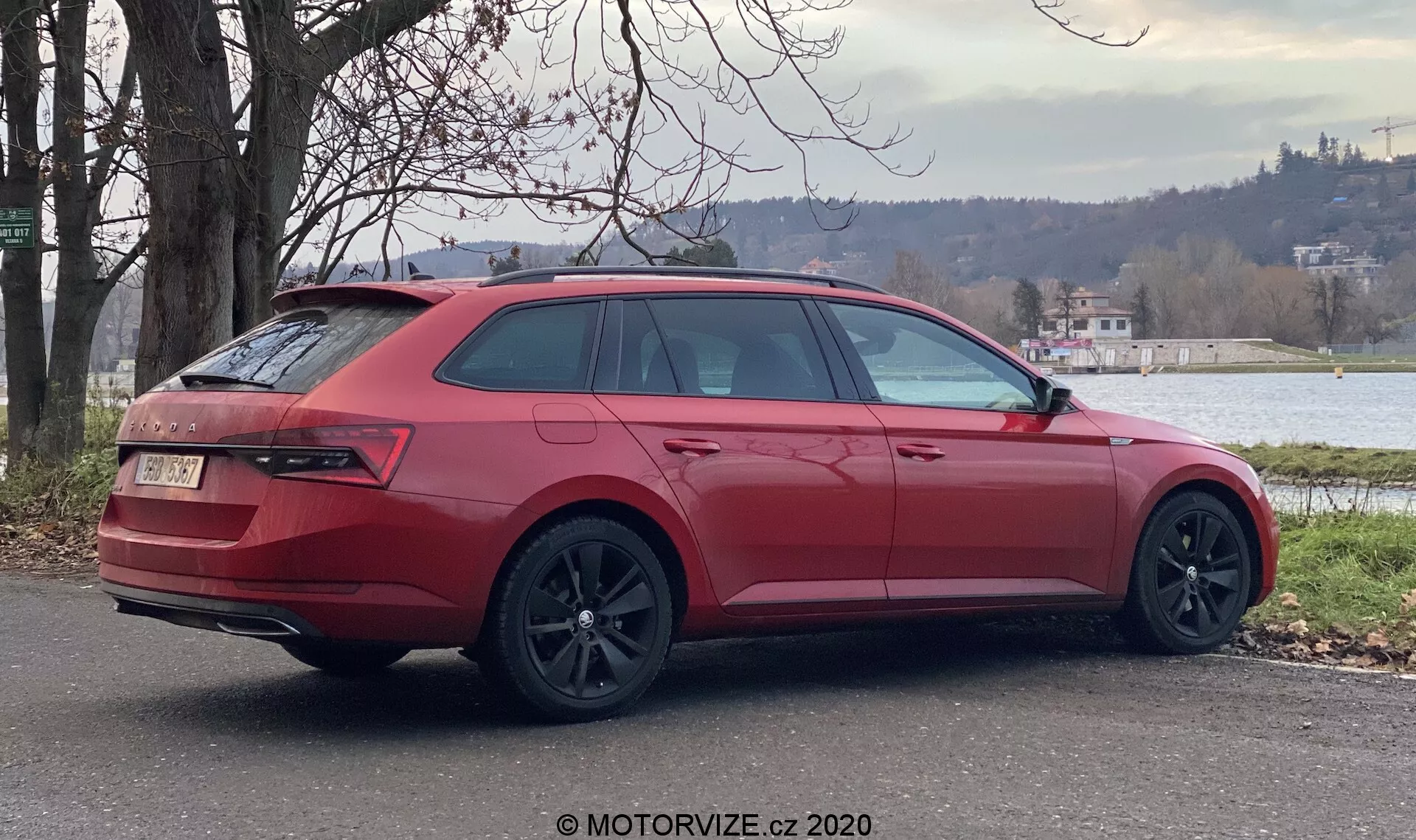 Červené kombi Škoda Superb 2019 zaparkované vonku, videné zo zadného rohového uhla, ktorý ukazuje pravú stranu vozidla. Kľúčové dizajnové prvky zahŕňajú veľké tmavé zliatinové kolesá, zvažujúce zadné a bočné okná, obklopujúce zadné svetlá, zatvorenú zadnú časť s odznakom, zadný nárazník s difúzorom a dvomi výfukmi, strešné lišty a farebne zladené bočné zrkadlo. Prostredie zahŕňa zeleň a vodnú plochu v pozadí, naznačujúc pokojné predmestské alebo polonovosídlené prostredie.
