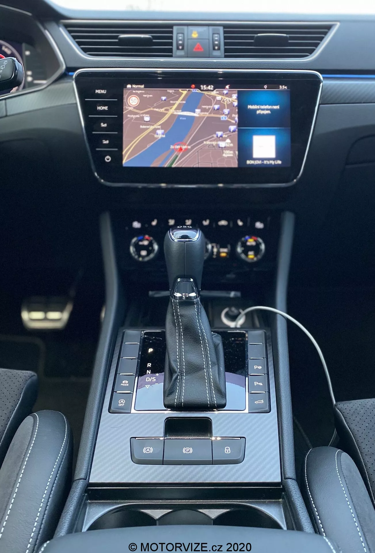 Pohľad na interiér modelu Škoda Superb Limousine z roku 2019, zameraný na stredovú konzolu a prístrojovú dosku z perspektívy zadného sedadla. Obsahuje automatickú radiacu páku s viditeľným symbolom 'P' pre parkovanie, tlačidlo elektronickej parkovacej brzdy, dotykové tlačidlá a dotykovú plochu. Voľne stojaci infotainment displej nad konzolou zobrazuje mapu a indikátor médií. Dvojzónová klimatizácia je umiestnená pod displejom a obklopená ventilačnými otvormi. Moderný, elegantný čierny interiér s chrómovými doplnkami a luxusnými prešívanými sedadlami. Viditeľná je horná časť multifunkčného volantu a časť prístrojovej dosky s analógovými ukazovateľmi. Nabíjací kábel pre smartphone naznačuje možnosti pripojenia.