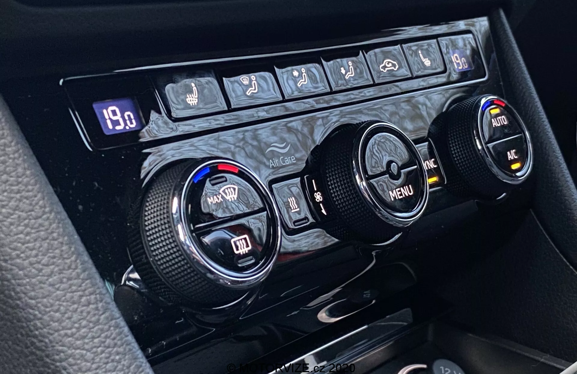 Detailný záber na klimatizáciu v Škoda Superb 2019 umiestnenú na stredovej konzole, s tromi kruhovými ovládačmi pre nastavenie teploty, navigáciu v menu a ďalšie funkcie. Ľavý ovládač reguluje teplotu pre vodiča, pravý pre spolujazdca, zatiaľ čo stredný ovládač ponúka navigáciu v menu so smerovými šípkami. Texturované okraje zabezpečujú ergonomické používanie. Nad ovládačmi je rad tlačidiel zahŕňajúci ovládanie zadného odmrazovania, predného odmrazovania, recirkulácie vzduchu, automatickej klimatizácie, klimatizácie a možno aj tlačidla varovania. Digitálny displej ukazuje '19.0,' čo môže indikovať nastavenú teplotu alebo inú hodnotu súvisiacou s prevádzkou systému.