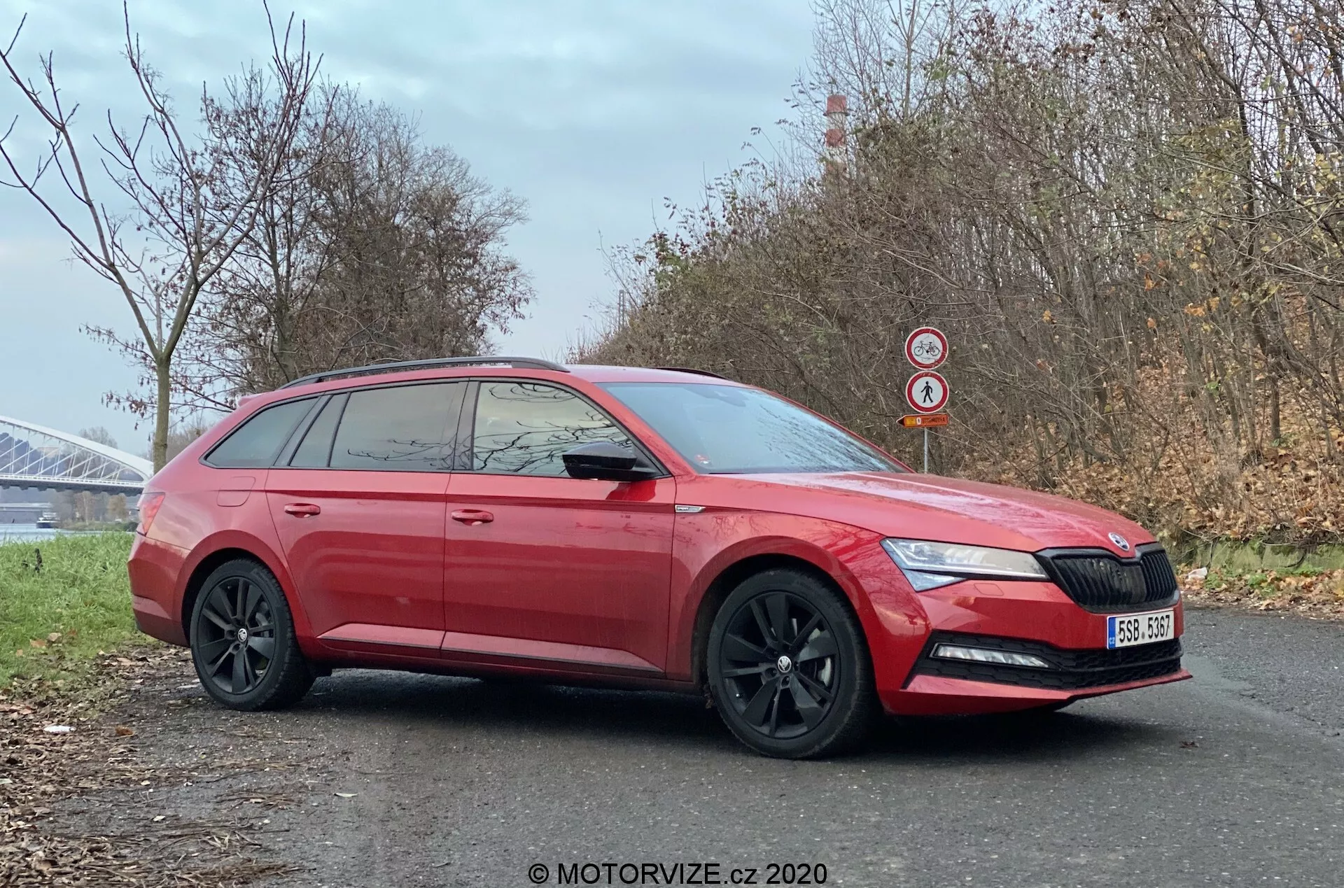 Bočný profil červeného kombi Skoda Superb z roku 2019 zaparkovaného pri ceste, ktorý zdôrazňuje jeho moderný dizajn s ostrými prednými svetlami vrátane denných LED svetiel, výraznou prednou mriežkou s logom, čiernymi zliatinovými kolesami, tónovanými oknami na zabezpečenie súkromia a predĺženou líniou strechy, typickou pre karosériu kombi. Fotografia, ktorá zvýrazňuje bok aj mierne skrátenú prednú časť, zachytáva detaily ako integrované smerovky v bočných zrkadlách, kľučky dverí v farbe karosérie, ochranné spodné obloženie a zadný strešný spojler. Čistý obraz zhotovený počas chladného obdobia pri možnej konštrukcii mosta umožňuje viditeľnosť vlastností auta a okolia.