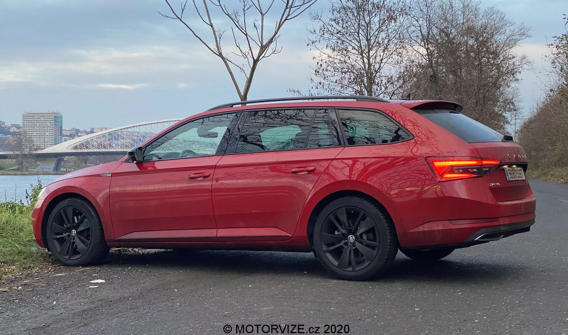 Obrázok zobrazuje moderný, živo červený Škoda Superb 2019 kombi z pohľadu zadnej tretiny. Auto je zaparkované na dláždenej ploche s riekom a mostom v pozadí, demonštruje štíhle línie, klesajúcu strechu vzadu, ľahko tónované okná s chrómovaným lemovaním a integrovaný zadný spojler. Športový dizajn je zdôraznený veľkými čiernymi zliatinovými kolesami s viditeľnými brzdovými strmeňmi, kontrastnými čiernymi zrkadlami, zapustenými kľučkami dverí, LED zadnými svetlami a parkovacími senzormi na zadnom nárazníku, čo naznačuje vyššiu úroveň výbavy s výkonom a pokročilou asistenčnou technológiou pre vodičov.