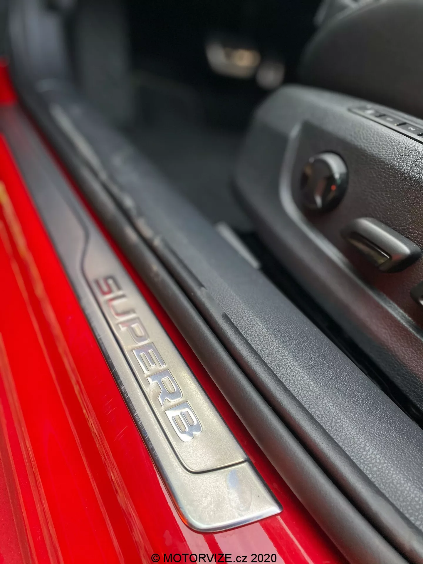 Detailný záber prahu dverí na strane spolujazdca vo vozidle Škoda Superb 2019. Obrázok zachytáva naklonený uhol zobrazenia nápisu 'SUPERB' na prahu z kartáčovanej nerezovej ocele, osvetlený pre lepší estetický dojem. Červený lak exteriéru kontrastuje s tmavým interiérom. Viditeľný okraj dverí s plastovým obložením a ovládaním elektrických okien zdôrazňuje pokročilé technológie. Plytká hĺbka ostrosti zvýrazňuje prah, čím vytvára bokeh efekt pozadia.