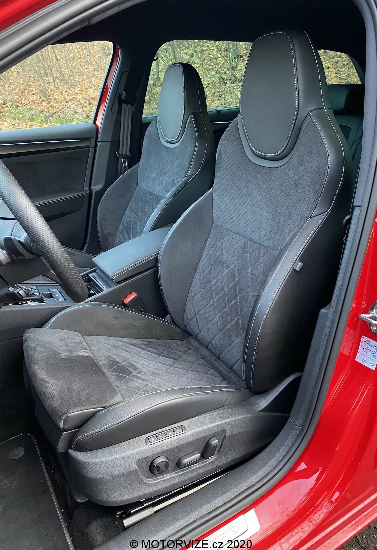 Interiér vozidla Škoda Superb 2019 so zameraním na sedadlo vodiča. Športové sedadlo s čalúnením z kože a materiálu podobného semišu, s prešívaním, integrovanou opierkou hlavy, výraznými opornými kontúrami a elektronickými ovládacími prvkami naznačuje výkonnostný dizajn, luxus a pokročilé komfortné funkcie. Viditeľný červený rám dverí naznačuje, že exteriér môže byť červený.