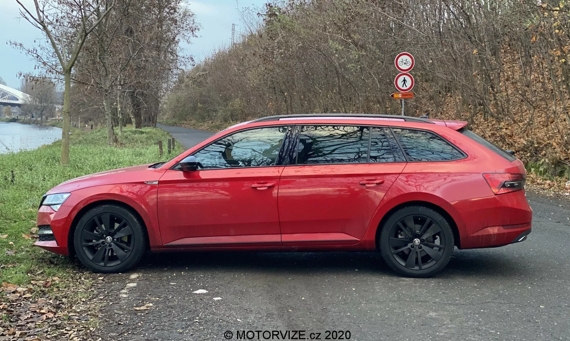 Bočný profil moderného kombi Škoda Superb (Superb-Limousine) z roku 2019 v červenej farbe, s predĺženou líniou strechy, čiernymi hliníkovými kolesami a farebne zladenými spätnými zrkadlami s integrovanými smerovkami. Auto stojí na asfaltovej ceste vedľa trávnatej plochy, za ktorou sú stromy a vodná plocha, za denného svetla.
