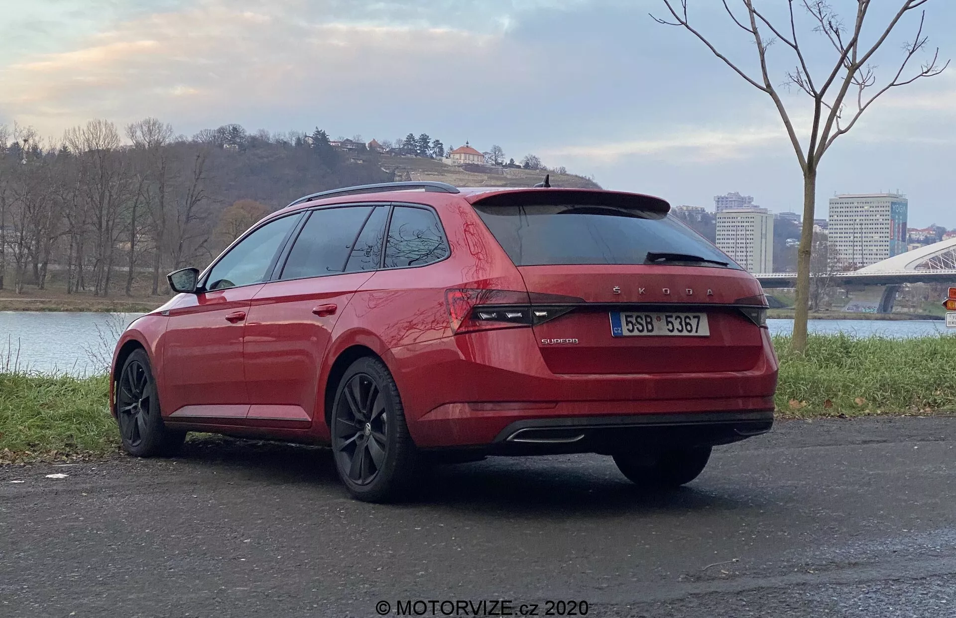Šikmý zadný pohľad na červený kombi Škoda Superb 2019 zaparkovaný vonku, zameriavajúci sa na zadnú a pravú stranu vozidla. Vyznačuje sa neprerušovanými zadnými svetlami cez dvere kufra, dvojitými výfukmi, tónovanými sklami, tmavými zliatinovými kolesami a nachádza sa na dláždenej ploche s vodnou plochou a mostom v pozadí.