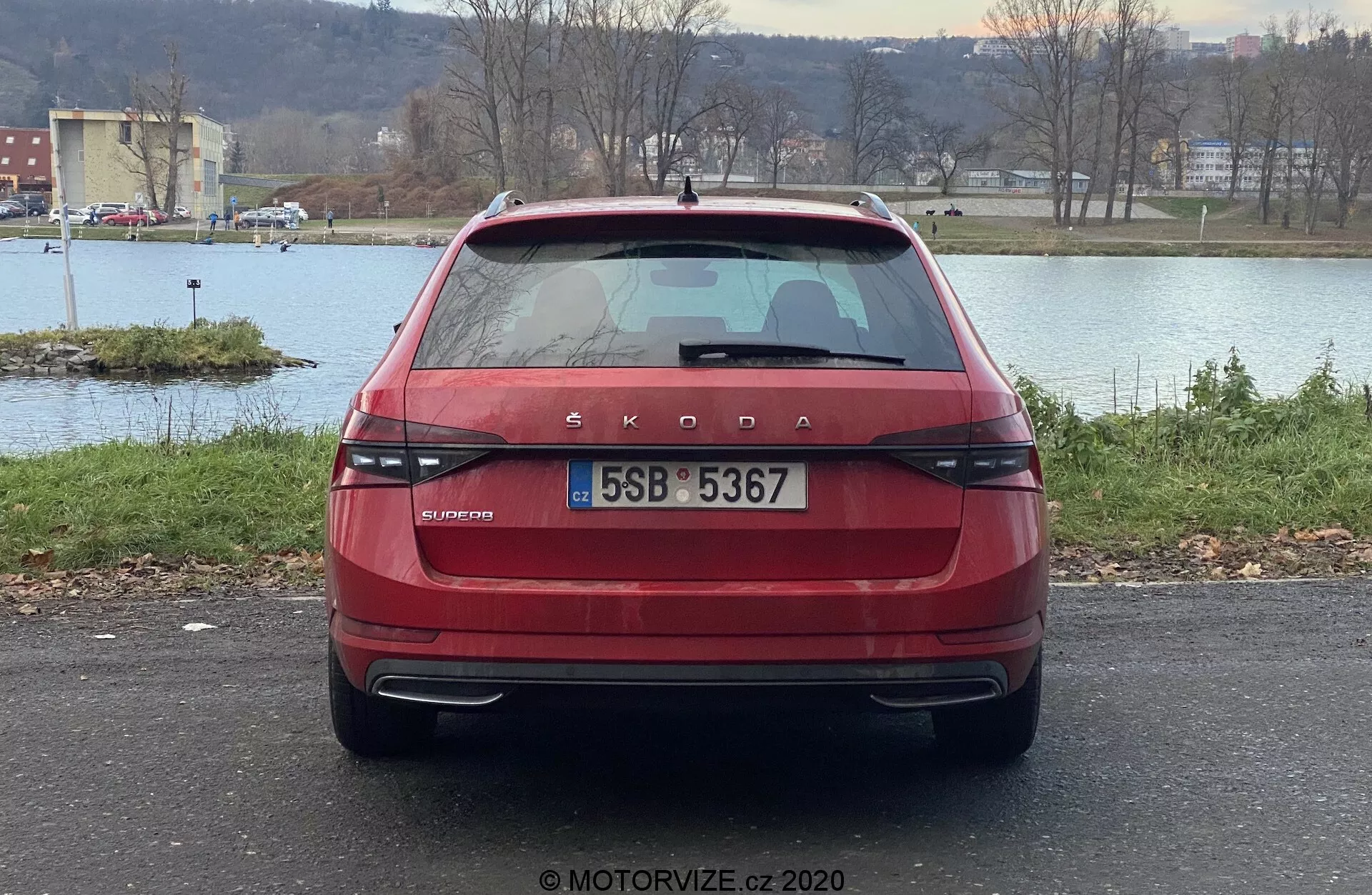 Zadný pohľad na červený Škoda Superb 2019 limuzínu, ukazujúci charakteristické značkovanie Škoda a nápis modelu OCTAVIA na kufri. Obrázok zvýrazňuje moderné, horizontálne usporiadané zadné svetlá, centrálne umiestnenú ŠPZ, zadný stierač, tónované zadné okno s odmrazovacími čiarami, vysoko umiestnené brzdové svetlo, parkovacie senzory na nárazníku a odrazky. Auto je zaparkované vonku s vodnou plochou, stromami a panorámou v pozadí.