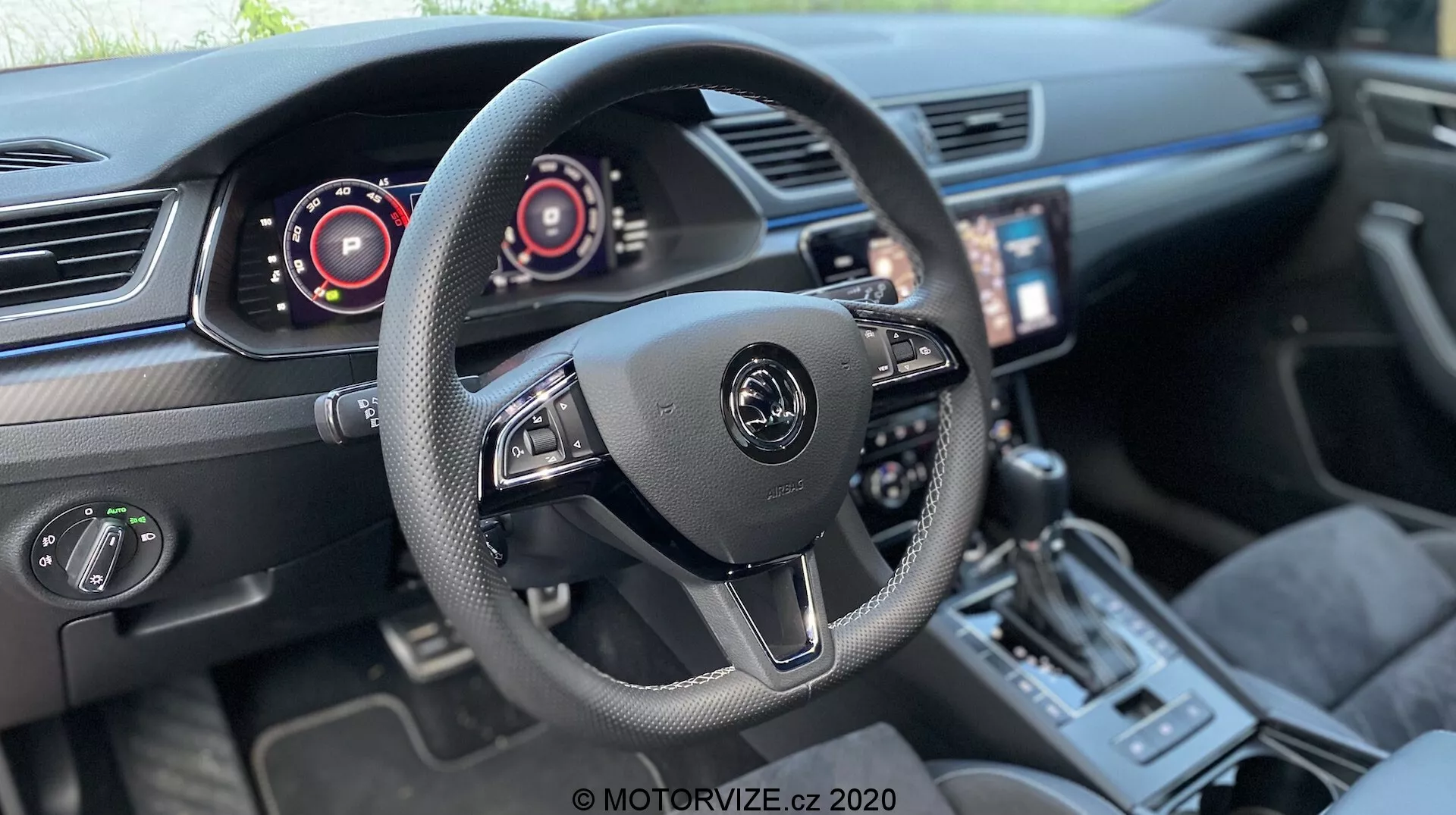 Interiér vozidla Škoda Superb Limousine 2019 zameraný na oblasť kokpitu vodiča. Centrálne umiestnený trojramenný volant s logom výrobcu a ovládacími prvkami pre audio a palubné systémy vrátane pádiel za volantom. Digitálne prístrojové panely za volantom naznačujú tachometer a rýchlomer. Dotykový infotainment displej naklonený smerom k vodičovi, dvojzónová klimatizácia, športový radič, prepínač jazdných režimov a kvalitné kožené sedadlá s kontrastným prešívaním naznačujú moderné, vysokovýbavené vozidlo s pokročilou technológiou.