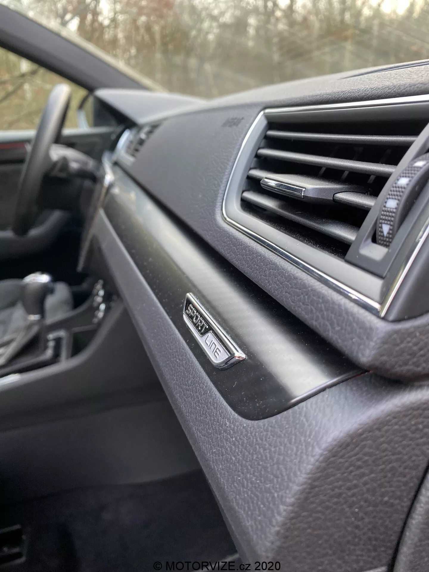 Detail interiéru Škoda Superb 2019, palubná doska z pohľadu spolujazdca. Zamerané na odznak 'Superb' pri vetracom otvore, moderný dizajn s mäkkým povrchom. Rozmazaný volant v pozadí, tmavá farebná schéma.