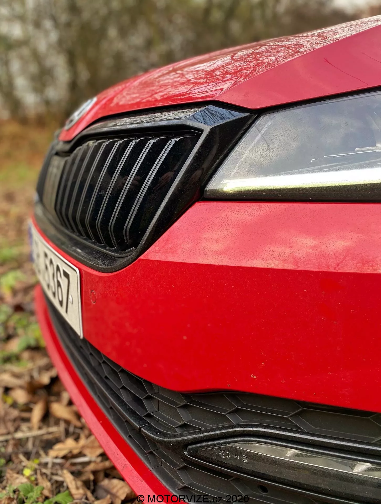 Detailný záber na prednú ľavú časť červeného vozidla Škoda Superb Limousine 2019. Obrázok zachytáva moderný dizajn pravého svetlometu s LED denným osvetlením, čiernu mriežku s vertikálnymi lamelami, aerodynamický okraj červeného kapotu, športový predný nárazník s veľkým nasávacím otvorom s horizontálnymi lamelami a priestor pre znak výrobcu, čiastočne zahalený. ŠPZ je mierne viditeľná, ale nerozostrená kvôli malej hĺbke ostrosti, s rozmazaným prirodzeným vonkajším pozadím.