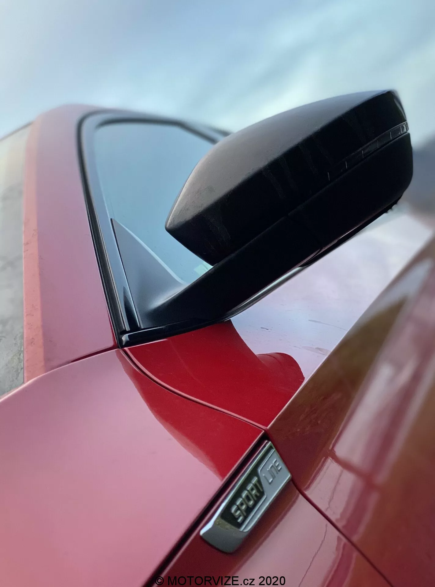 Detailný pohľad na bočné spätné zrkadlo na žiarivo červenom vozidle Skoda Superb 2019 limuzína, zvýrazňujúci čierne aerodynamické puzdro zrkadla a LED bočné smerovky pod ním, s viditeľnou časťou stĺpika vozidla, zachytené z nízkeho uhla, ktorý zvýrazňuje čistý dizajn a lesklý povrch, označený vodoznakom MOTORVIZE.cz 2020.