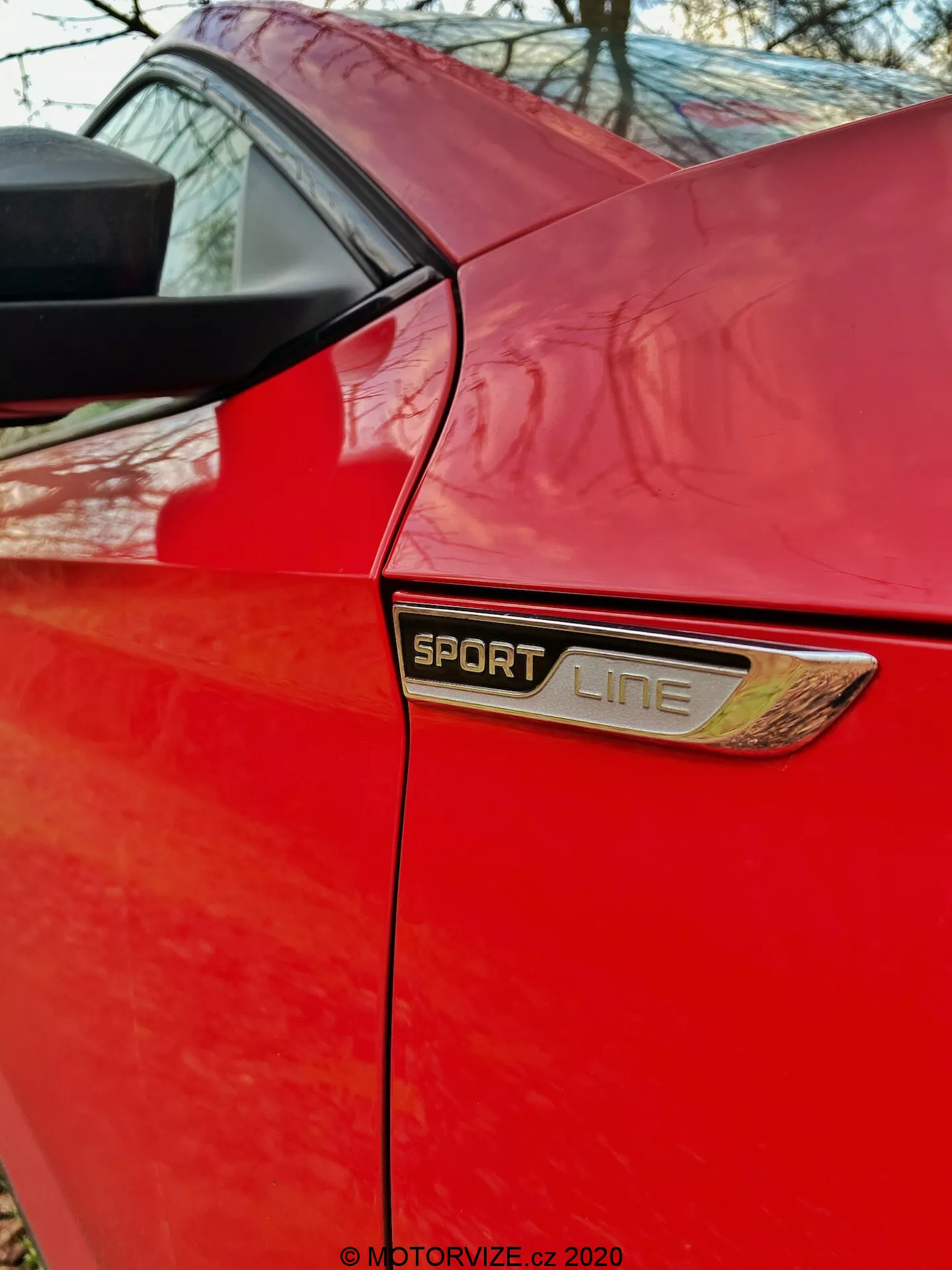 Detailný záber bočnej časti zadného štvrťpanelu červenej Škody Superb 2019 limuzíny. Tento obrázok sa zameriava na oblasť za bočnými dverami, kde je prominentný 'SPORT LINE' odznak s chrómovým alebo kovovým rámom a prídavným ozdobným vetracím dielcom pod ním. Nad odznakom je viditeľný spodný okraj zadného bočného okna a zadný stĺpik podporujúci štruktúru strechy. Fotografia zdôrazňuje špecifické úrovne výbavy, estetické prvky a kvalitnú farbu športovej verzie