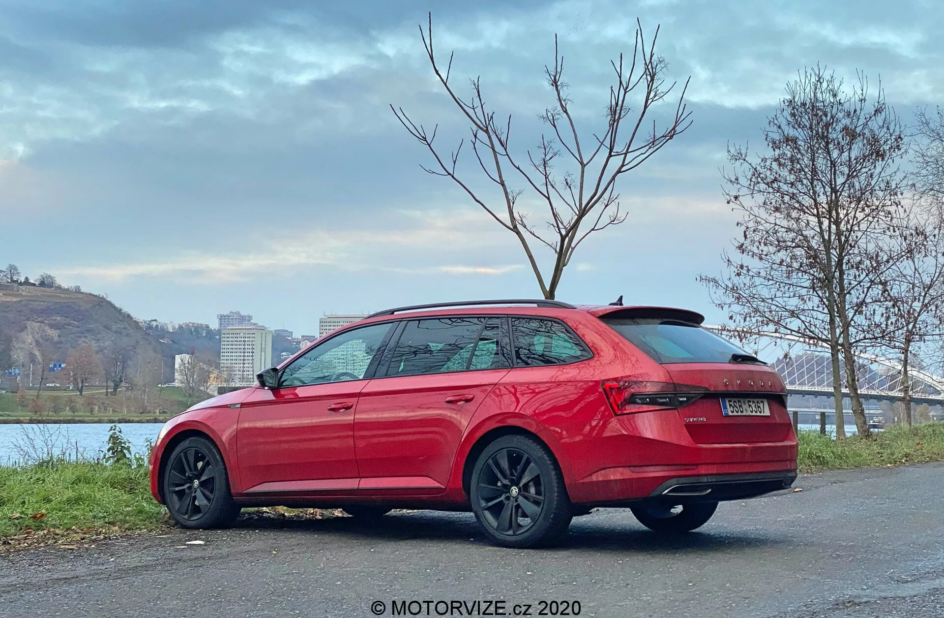 Obrázok vozidla Škoda Superb z roku 2019, kombi, pohľad zozadu tri štvrtiny, ukazuje vysoko umiestnené obklopujúce zadné svetlá, robustný nárazník, žiadne viditeľné výfukové rúry, strešný spojler, elegantné bočné kontúrové línie, dverové kľučky v farbe karosérie, ochranné spodné bočné prahy, chrómové okenné lišty, tmavé viacramenné kolesá, stojaci na dláždenom povrchu blízko brehu rieky s mostom a stromami, zatiahnutá obloha, vodoznak MOTORVIZE.cz 2020.