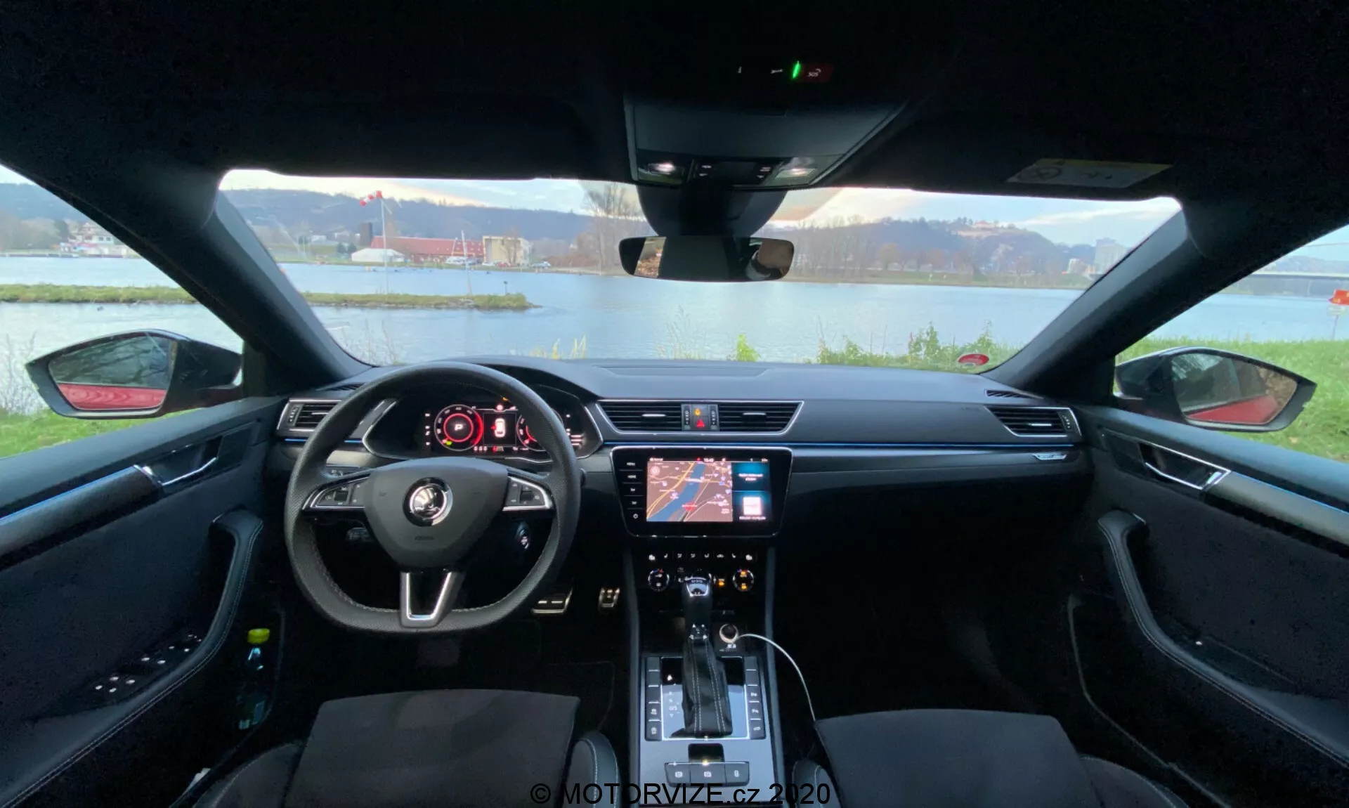 Pohľad na interiér Škoda Superb 2019 zo zadného sedadla smerom k prístrojovej doske, zachytávajúci šírku interiéru vozidla. Centrálne umiestnený volant s ovládacími prvkami a logom Škoda je výrazne viditeľný v popredí. Za volantom sa nachádza digitálny prístrojový displej ukazujúci dôležité informácie o vozidle. Stredová konzola obsahuje infotainment systém s navigáciou a nastaveniami médií, spolu s ovládačmi klimatizácie a funkčnými tlačidlami pod ním. Páková riadiaca páka, držiaky na nápoje a detailné sedadlá s kontrastným prešívaním zdôrazňujú moderný estetický vzhľad zameraný na pohodlie a komfort vodiča.