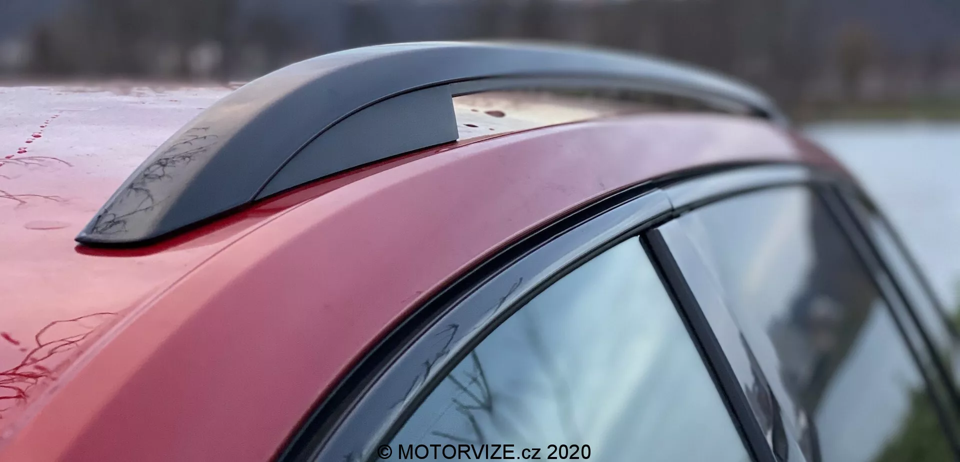 Detailný bočný pohľad na časť vozidla Škoda Superb 2019, zameraný na červenú strechu, bočný stĺpik a príslušenstvo v podobe antény tvaru žraločej plutvy, so zreteľnými kvapkami vody naznačujúcimi voskovaný alebo hydrofóbny povrch; aerodynamický dizajn vozidla je zrejmý z vytvarovania a sklonu strechy