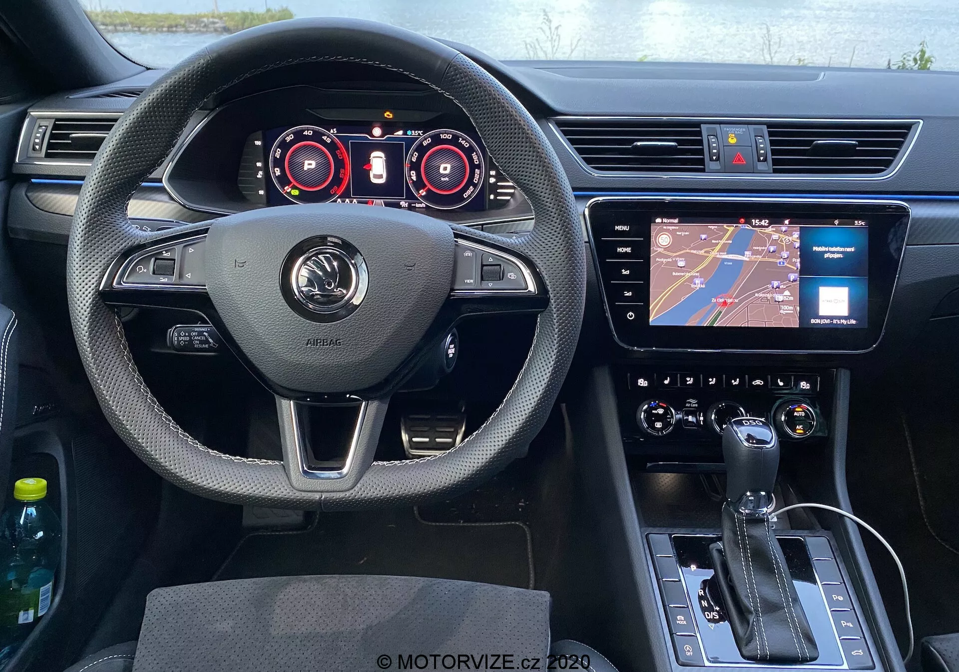 Pohľad z miesta vodiča na interiér vozidla Škoda Superb 2019 s centrálnym umiestnením volantu s opierkami na palce, multifunkčnými tlačidlami a výrazným logom výrobcu. Za volantom je digitálny prístrojový panel s ukazovateľmi rýchlomera a otáčkomera a s ďalšími displejmi pre prevádzkové údaje. Uprostred palubnej dosky je dotyková obrazovka infotainmentu s navigáciou, pod ňou je klimatizačný systém s tlačidlami a otočnými ovládačmi. V spodnej časti centrálnej konzoly je radiaca páka automatickej prevodovky, tlačidlo elektronickej parkovacej brzdy a ovládací koliesko. Vidieť aj časť dverí vodiča a spätné zrkadlo, s viditeľným ovládaním dverí. Kabína je kombináciou textúrovaných materiálov, chrómových prvkov, koženého volantu a lesklého čierneho plastu na konzole, čo dodáva moderný vzhľad.