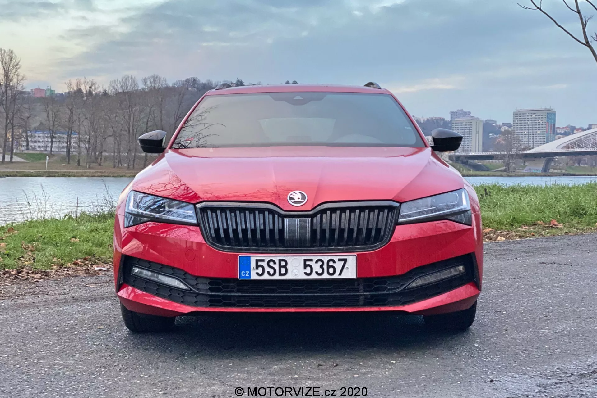 Predný pohľad na modernú červenú Škoda Superb Limousine 2019, ktorý ukazuje jej hladké aerodynamické kontúry, výraznú mriežku svislých lamiel s centrálnym výrobcom emblémom, štíhle predĺžené LED svetlomety, širokú mriežku na spodnom nárazníku s hmlovými svetlami, centrálnu ŠPZ a možný senzor alebo kameru pokročilých asistenčných systémov pre vodiča. Čelné sklo odráža oblohu, čo naznačuje senzor na stierače pri daždi alebo svetelný senzor pre automatické zapínanie svetlometov. Auto je zaparkované na dláždenej ploche s mestským pozadím, zdôrazňujúc jeho dizajn a vlastnosti.