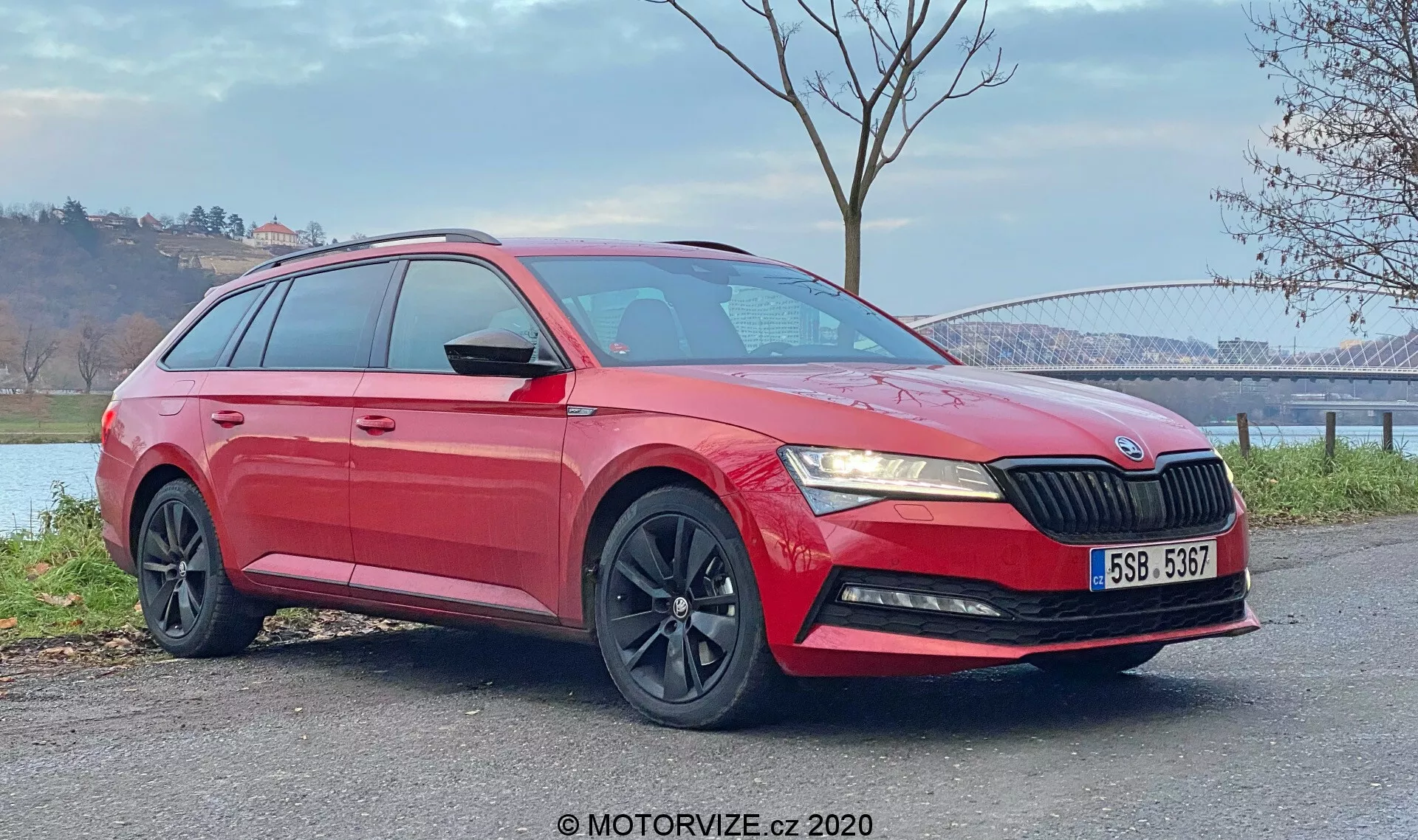 Obrázok červeného kombi Skoda Superb 2019 zachytený z trojštvrťového predného pohľadu. Fotografia zvýrazňuje prednú masku s vertikálnymi lamelami, elegantné uhlové svetlomety, hmlové svetlá v dolnom nárazníku, čierne viacramenné zliatinové kolesá a lesklý lak so zreteľnými charakterovými líniami a strešnými lištami. Viditeľné sú aj bočné zrkadlá lakované v farbe karosérie s integrovanými smerovkami a tónované okná v zadnej časti vozidla. Vozidlo je pozicované na rovnú povrch vonku a je vidieť s prírodným pozadím s mostom a vodnou plochou v diaľke. Vďaka dennému svetlu sú zreteľne viditeľné spodné aj horné časti vozidla.