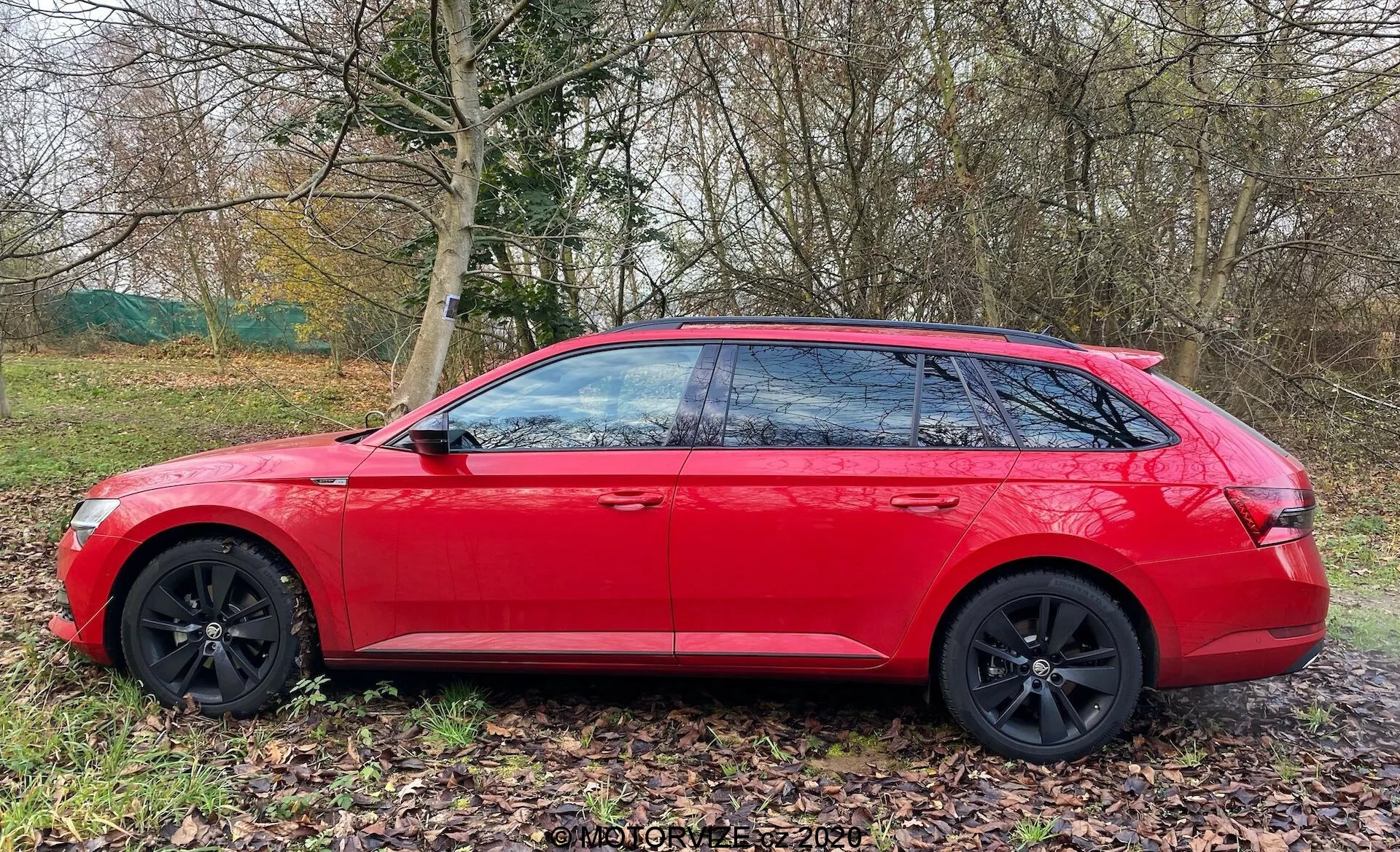 Škoda Superb kombi 2019 v jasne červenej farbe zobrazený z bočného pohľadu. Moderný dizajn sa vyznačuje ostrými geometrickými líniami, čiernymi viaclúčovými zliatinovými kolesami, tónovanými oknami s čiernymi B-stĺpikmi a klesajúcou strešnou líniou pre elegantný aerodynamický profil. Auto je zaparkované na trávnatej ploche vedľa cestičky, s prírodou v pozadí, čo naznačuje, že je vhodné na rodinné výlety alebo dobrodružstvá.