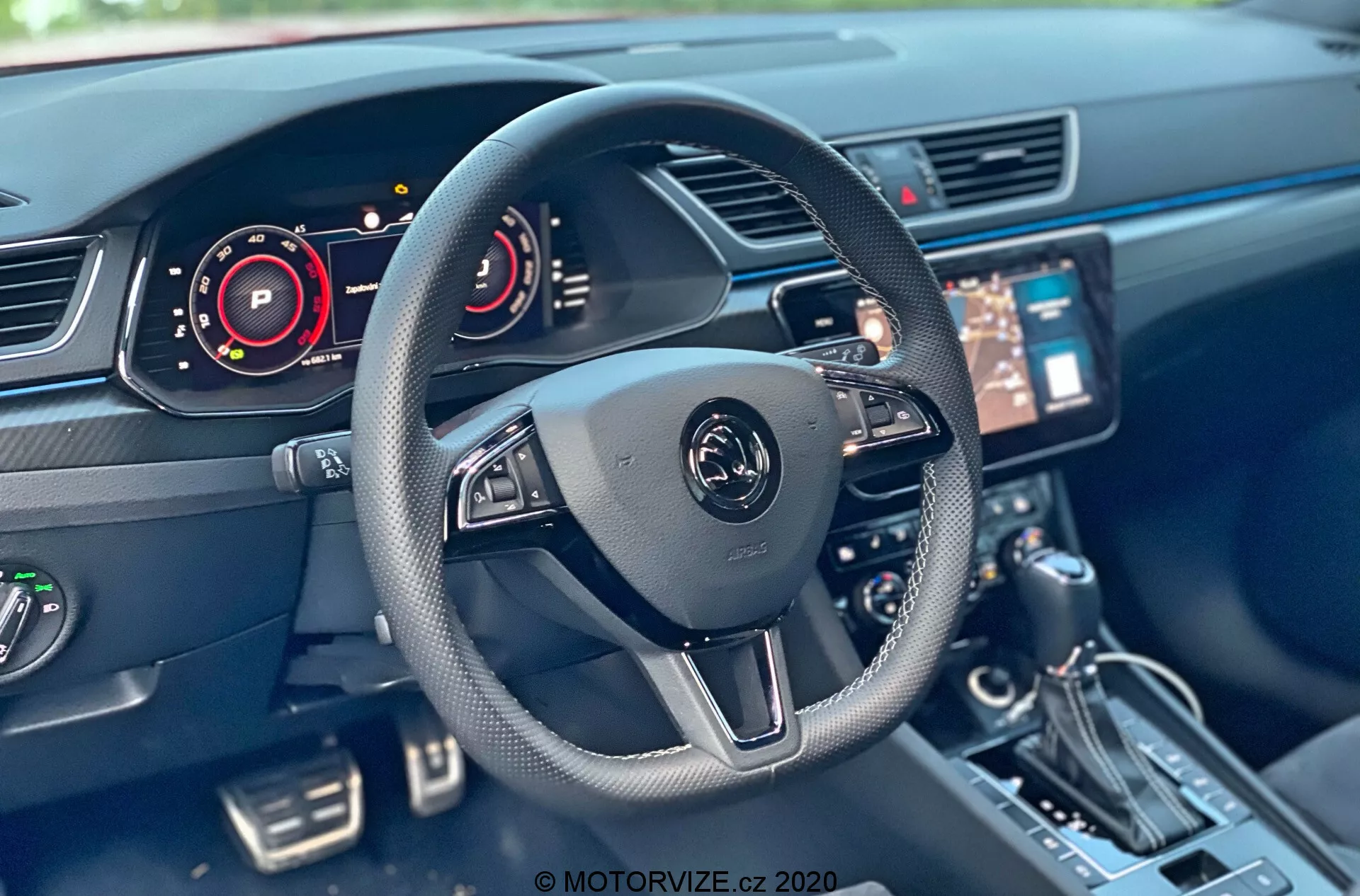 Interiér modelu Skoda Superb 2019 z pohľadu vodiča, s moderným trojramenným volantom s multifunkčnými ovládačmi a centrálnym emblémom. Viditeľné komponenty zahŕňajú digitálnu prístrojovú dosku, centrálny infotainment systém s dotykovou obrazovkou, konzolu, radiacu páku, tlačidlá a klimatizačný systém. Technológia interiéru vozidla ukazuje prvky ako digitálne prístrojové dosky a ovládanie volantu.