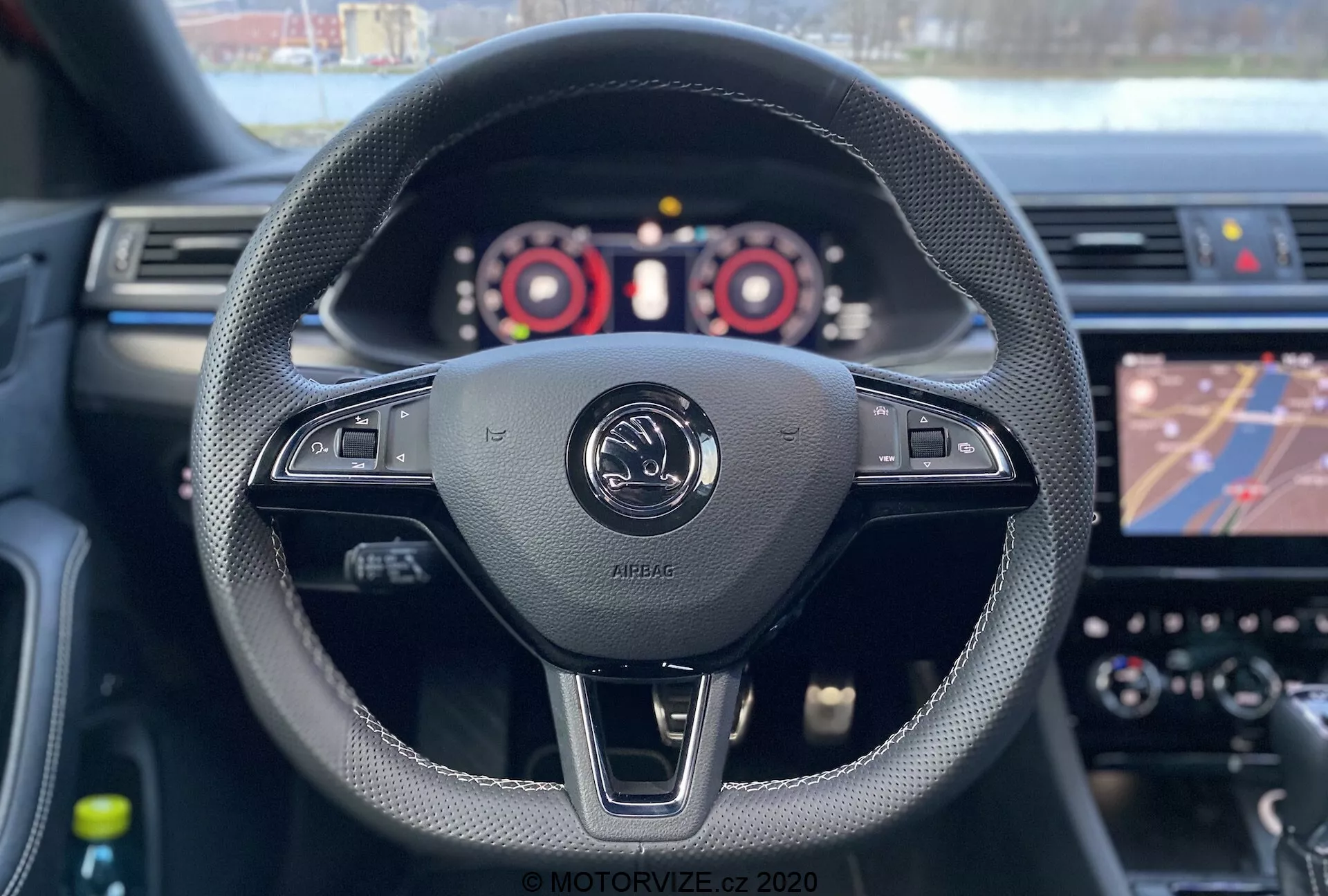 Pohľad na interiér Škoda Superb Limousine 2019 z pohľadu vodiča. Sedadlo vodiča odhaľuje volant ovinutý čiernou kožou s šitými detailmi, obsahujúci rôzne ovládacie prvky vrátane úpravy hlasitosti a pravdepodobných nastavení vozidla. Uprostred obrázka je emblém výrobcu s nápisom 'AIRBAG', zdôrazňujúci predný airbag pre vodiča. Viditeľné sú páky radenia, ktoré naznačujú ručné radenie. Prístrojový panel s tachometrom, rýchlomerom a digitálnym displejom je čiastočne viditeľný, zatiaľ čo infotainment systém zobrazuje navigačnú mapu. Plytká hĺbka ostrosti sa zameriava na volant a emblém, pričom prístrojová doska a dotykový displej sú mierne rozmazané.